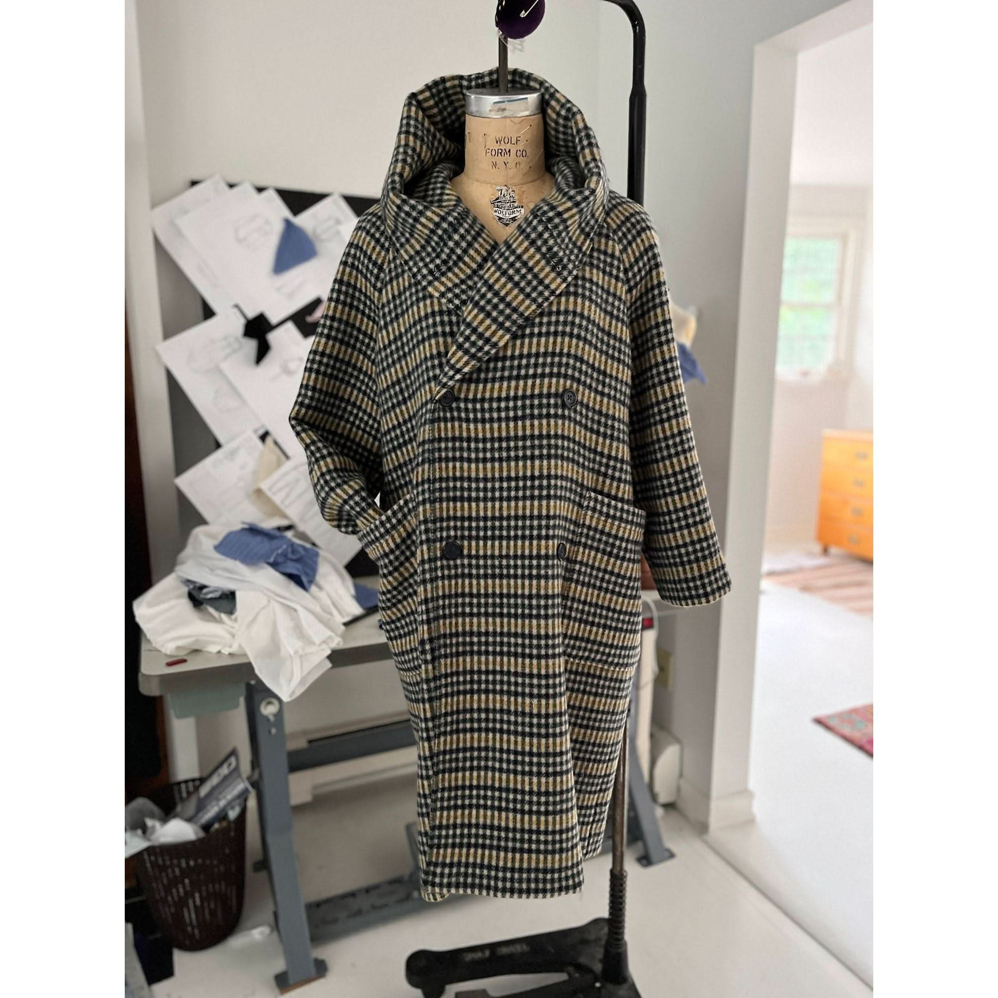 The Heather Harlan Hooded Blanket Coat, crafted from recycled Italian wool, is showcased on a dress form. The workspace around it is adorned with sewing patterns and fabric, creating the ambiance of a tailor or designer's studio. Its oversized draped hood enhances its stylish appeal.