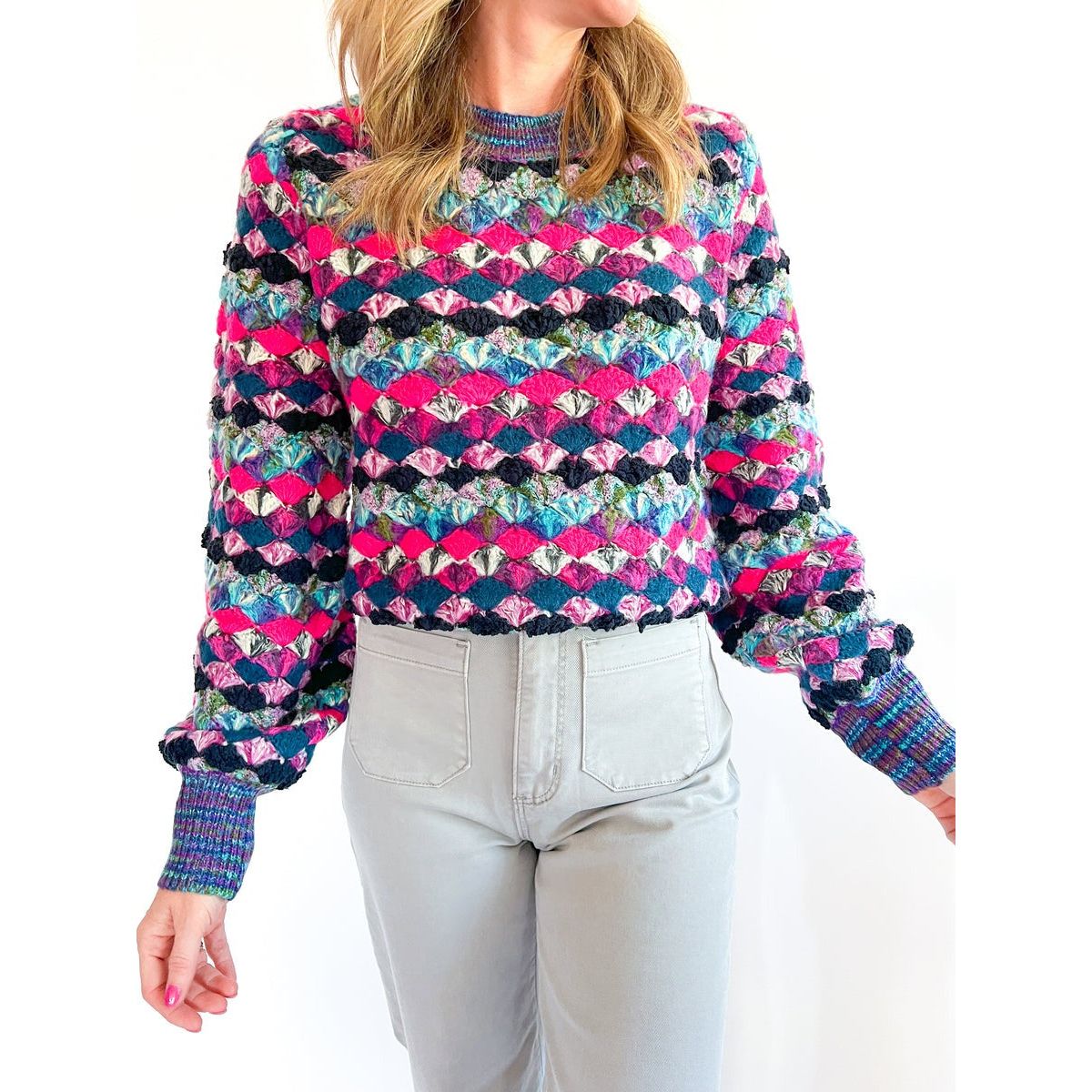 A person is wearing the stylish Allison New York Emerson Pullover, which boasts a vibrant pattern featuring various shades of pink, blue, and green. The pullover has textured detailing on its long, cuffed sleeves and is adorned with elegant crystal buttons. The individual pairs this eye-catching piece with light gray pants against a plain white background.