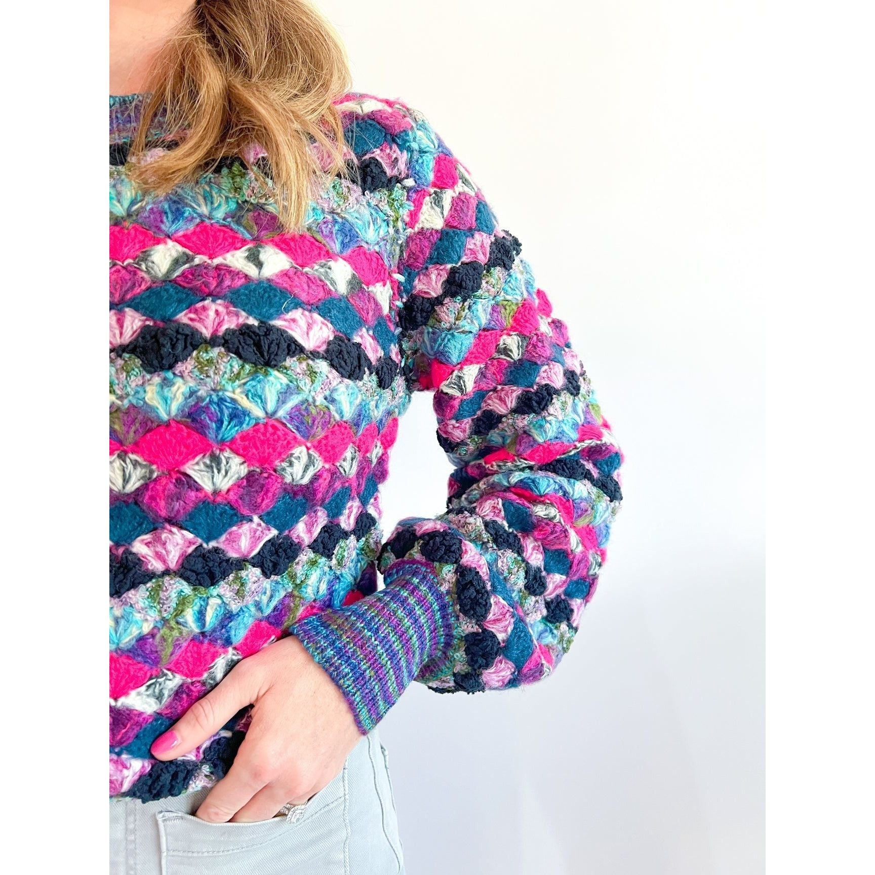 A close-up of an individual wearing a vibrant Allison New York EMERSON PULLOVER by Allison, featuring textured, geometric patterns in shades of pink, blue, purple, and green. Only the upper body is visible, with one hand resting on the hip and light-colored pants partially visible. The hot pink fuzzy yarns add a playful touch against the plain white background.