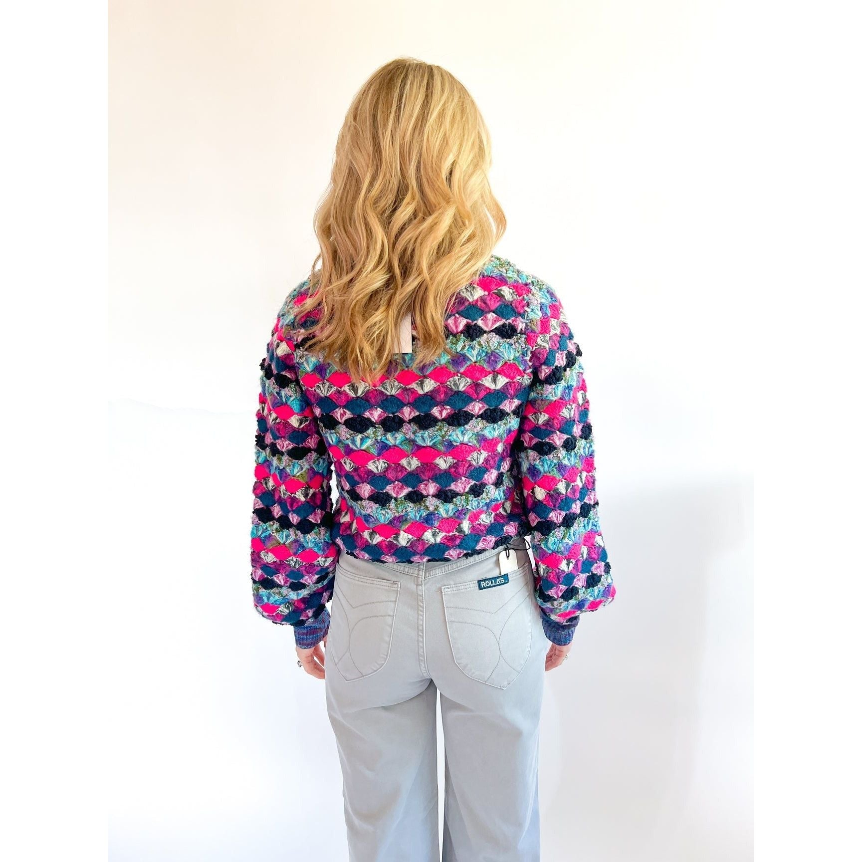 A person with long, wavy blonde hair stands facing away from the camera. They're wearing the Allison New York EMERSON PULLOVER by Allison, a colorful, patterned sweater featuring shades of blue, hot pink, and purple adorned with crystal buttons, paired with light grey pants. The background is plain white.