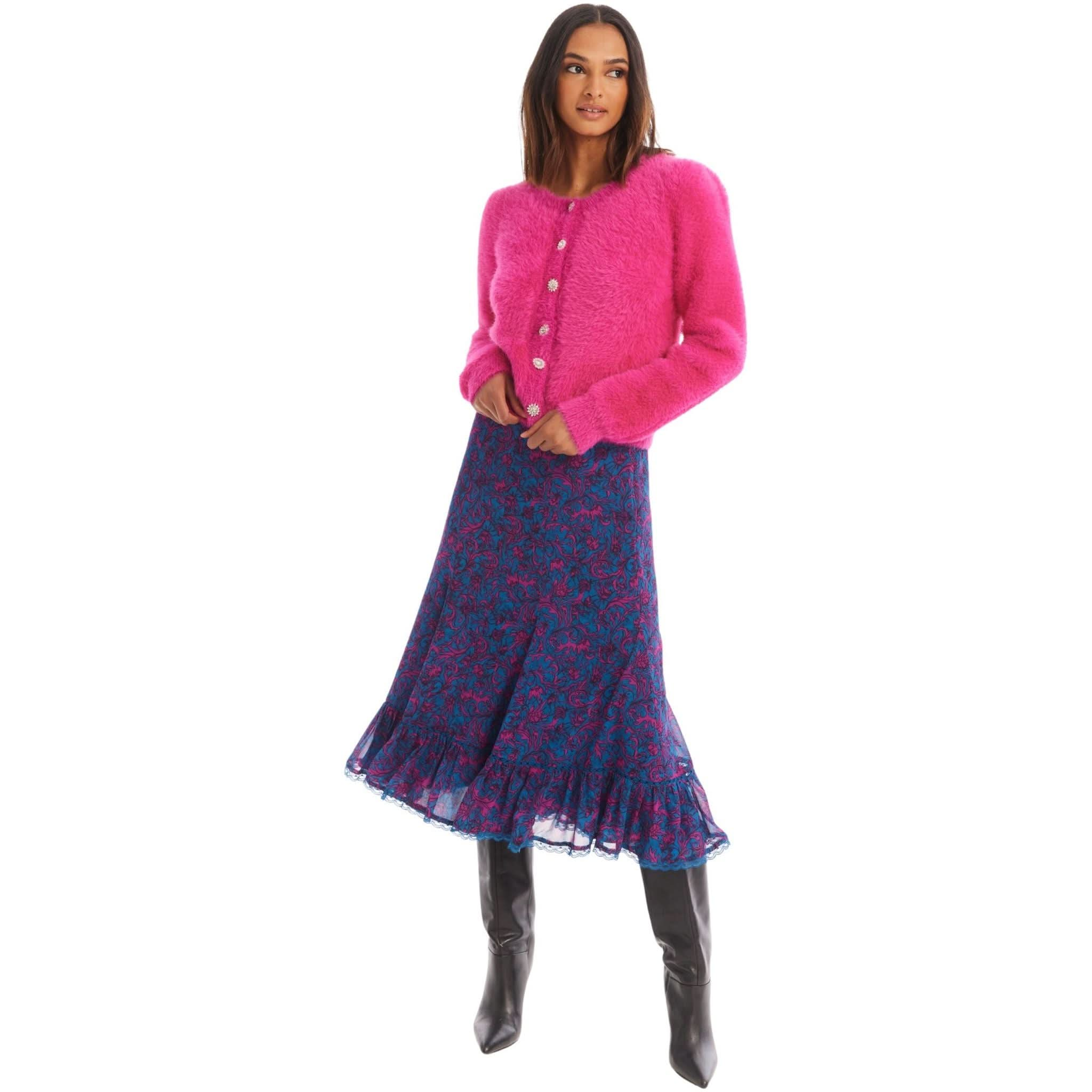 A woman wearing a bright pink, fuzzy, long-sleeve Allison New York Jeweled Sloan Cardigan by Allison with pearl buttons is paired with a navy blue and pink floral skirt that flares at the hem. She completes the look with black knee-high boots, standing against a white background.