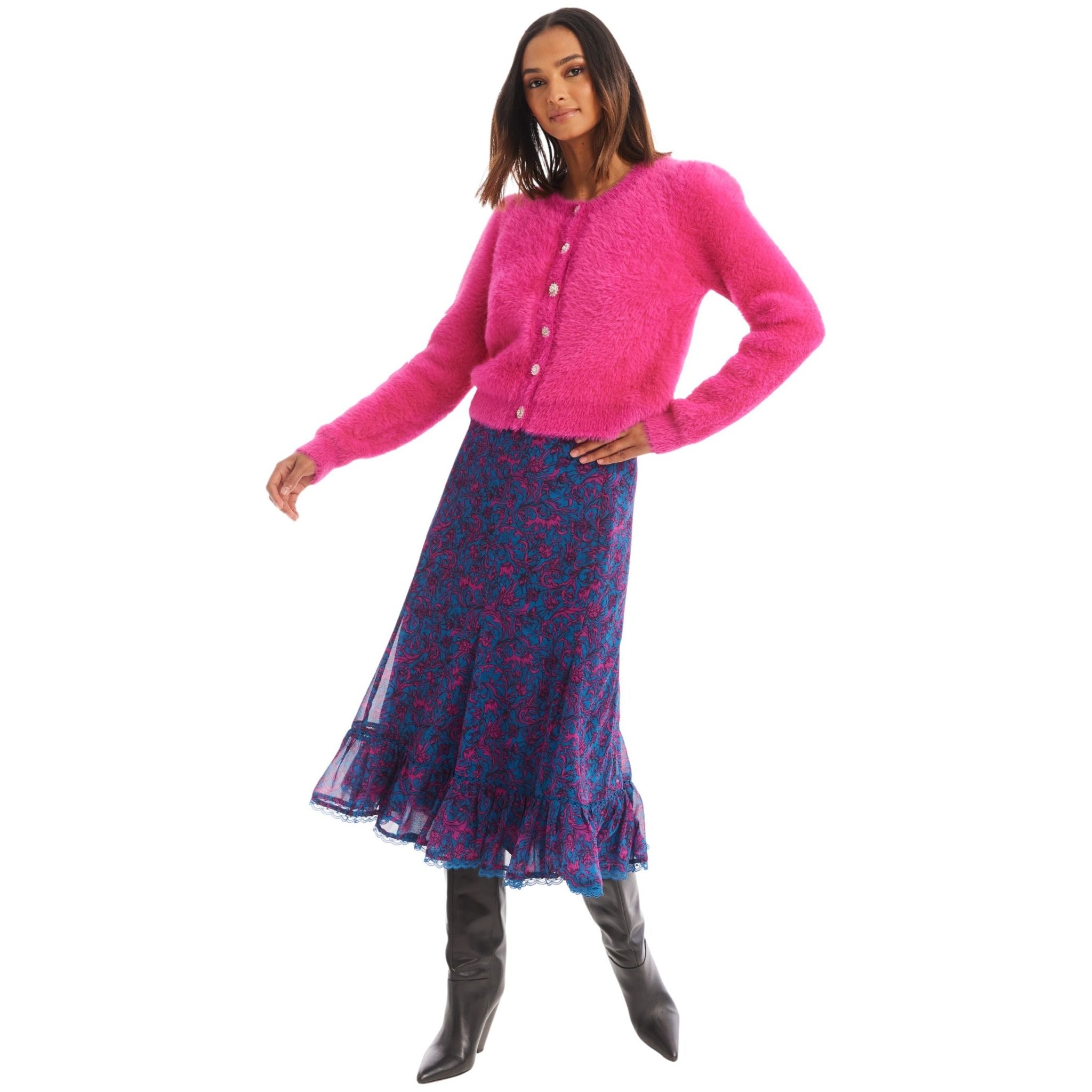 A woman stands modeling a bright pink Allison New York Jeweled Sloan Cardigan, made of lightweight viscose yarns and adorned with pearl buttons. It's paired with a blue and magenta-patterned midi skirt featuring a ruffled hem. She completes the look with black knee-high boots, her right hand on her hip as she gazes off to the side.