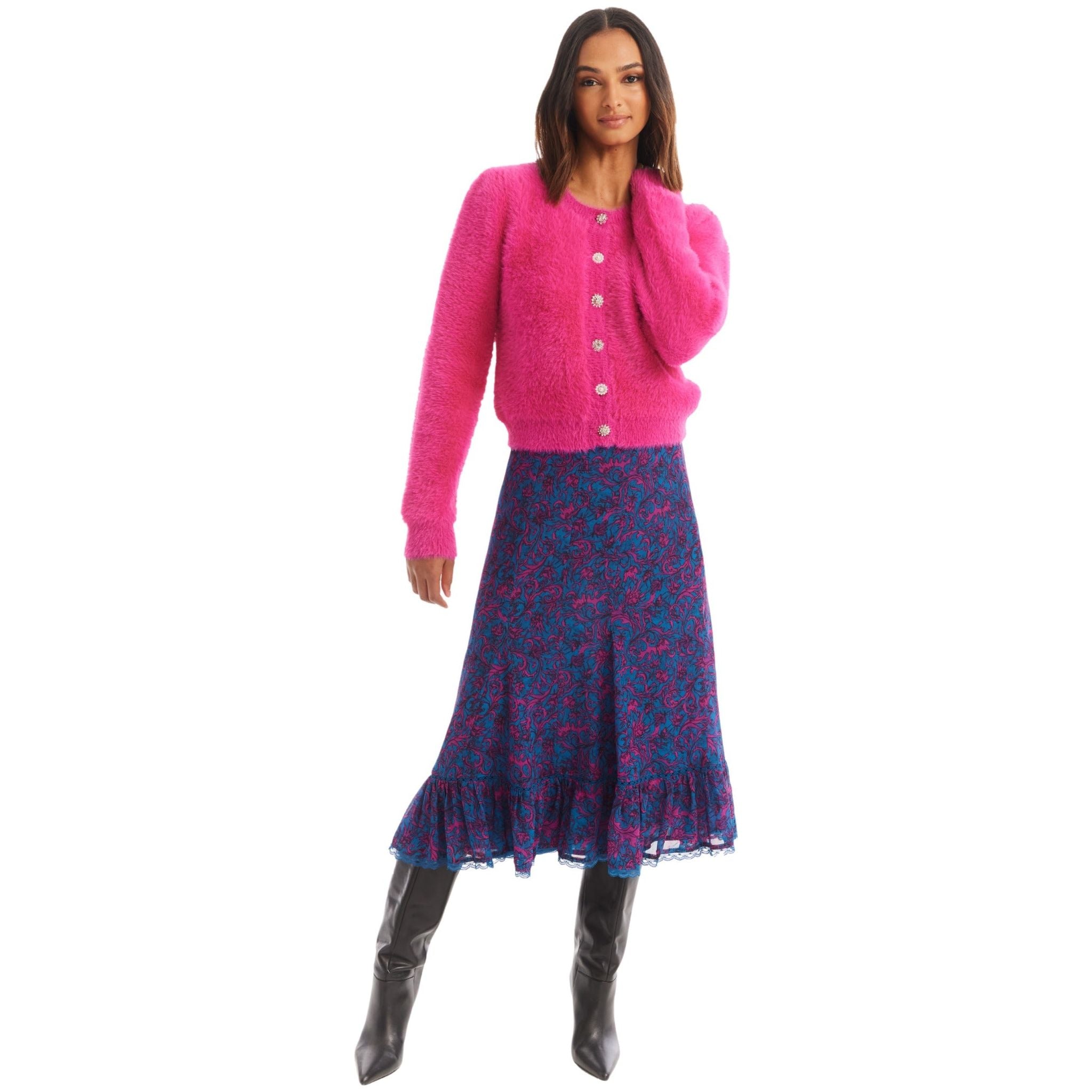 A woman stands with her left hand touching her hair, wearing an Allison New York JEWELED SLOAN CARDIGAN made of lightweight viscose yarns. She pairs it with a blue ankle-length skirt featuring a colorful floral pattern and black knee-high boots. She has a confident expression and stands against a plain white background.