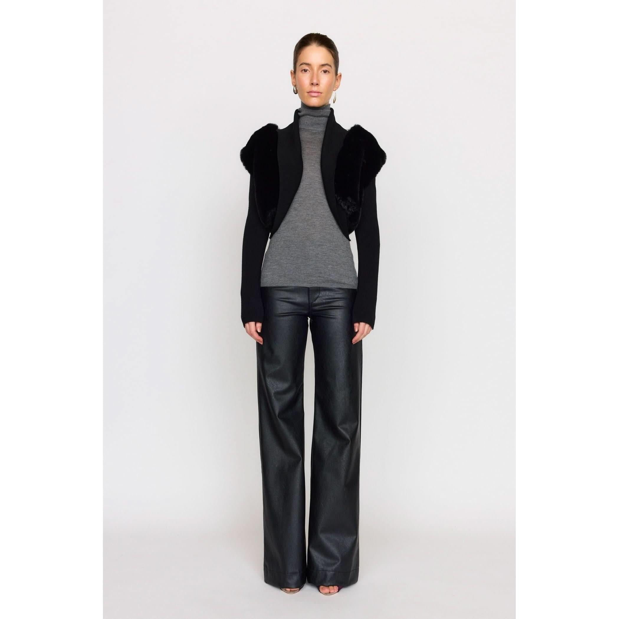 A person stands against a white background, styled in the Christy Lynn Criselle Knit in black over a gray turtleneck and paired with black wide-leg vegan leather pants. Their arms are relaxed by their sides as they face the camera, exuding effortless style.