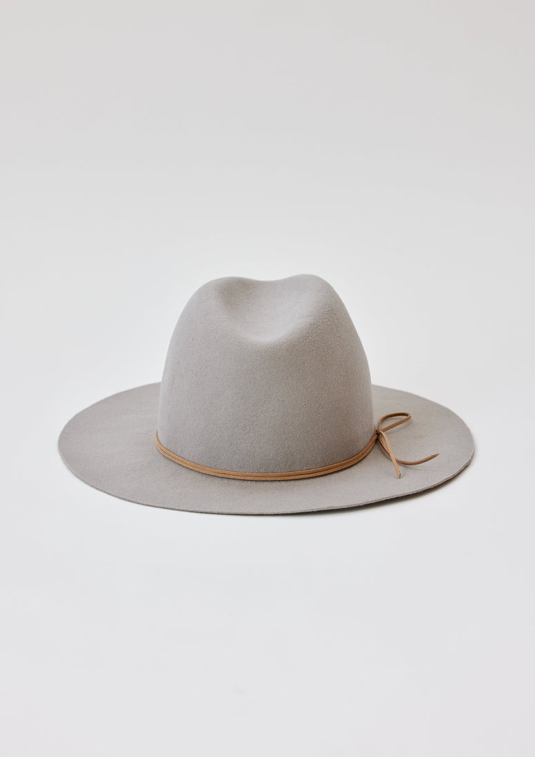 A Hat Attack Classic Amelia in light grey is displayed against a white background. This cotton denim, wide-brimmed fedora hat boasts a sleek brown leather band adorned with a small bow on the right side. The indented crown provides the piece with a structured and timeless look.