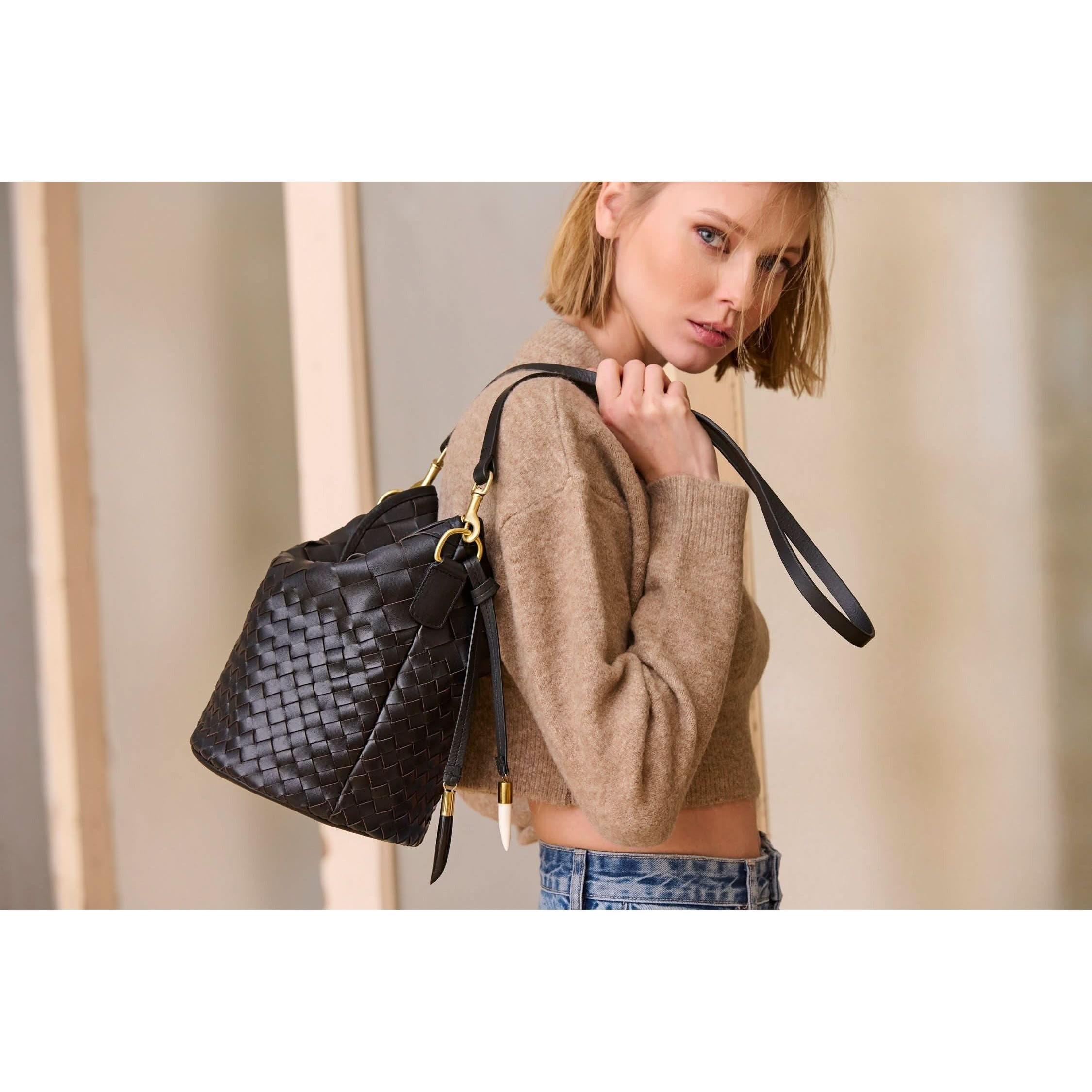 A person with shoulder-length blonde hair and a neutral expression holds a Kempton & Co Richmond Bucket Bag - Black Threaded Weave over their shoulder. They are wearing a cropped, beige sweater and blue jeans. The background is blurred with light-colored vertical elements.