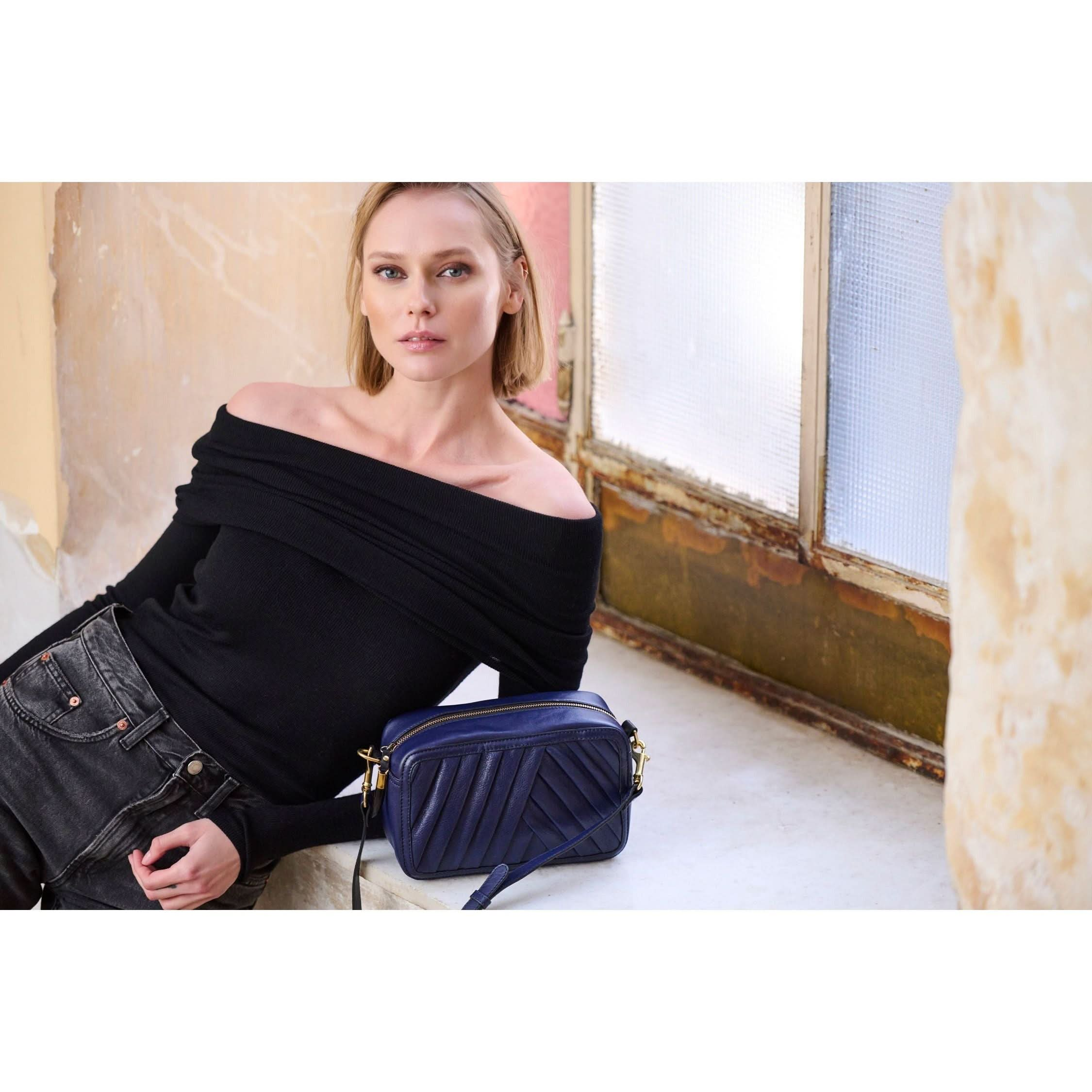 A woman with blonde hair is leaning against a windowsill, wearing a black off-shoulder top and dark jeans. She is holding the Kempton & Co Amberleigh Crossbody Bag in Pleated Indigo, showcasing the indigo hue that's in vogue. The background shows an aged wall and a textured glass window, creating an atmosphere of timeless elegance.