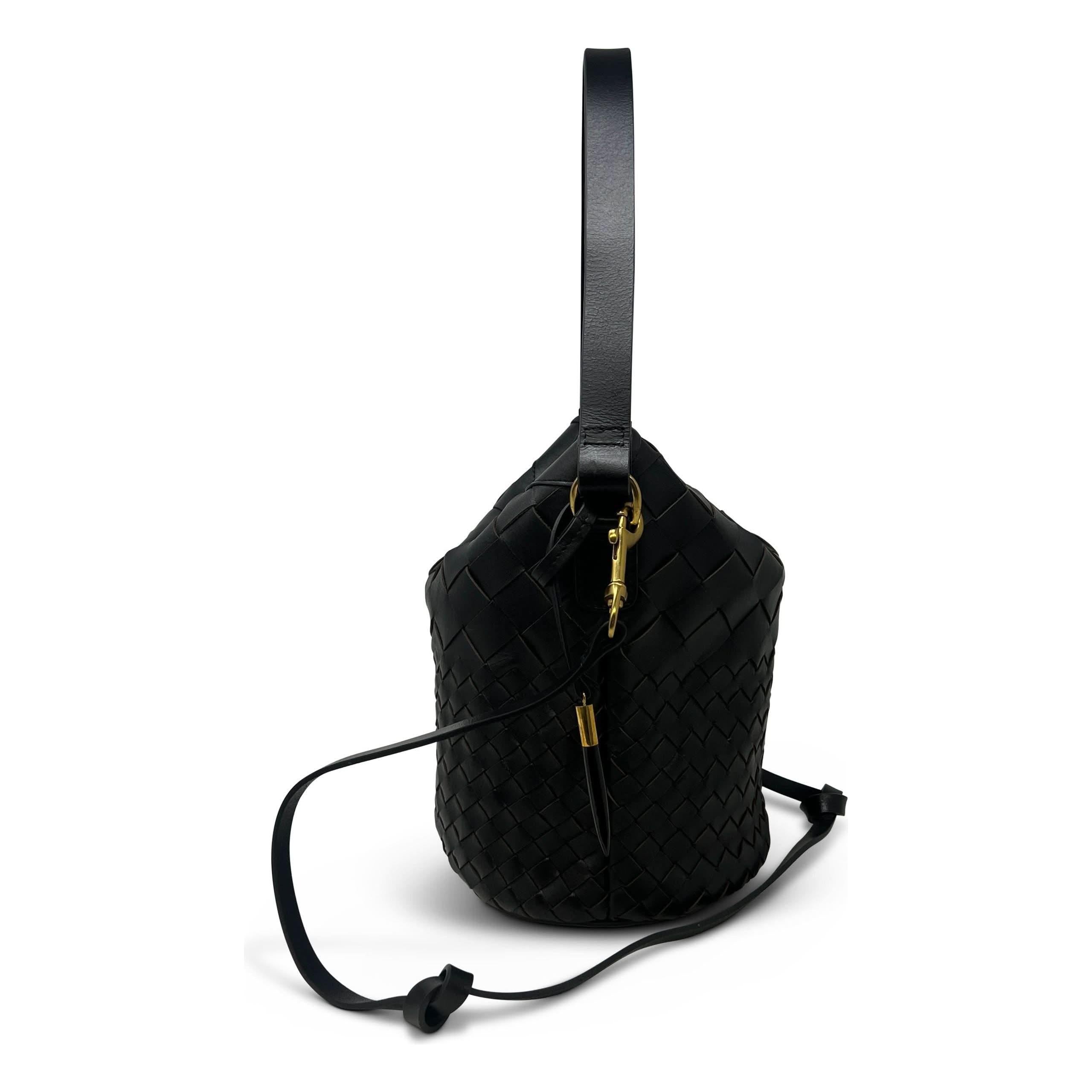 The Kempton & Co Richmond Bucket Bag - Black Threaded Weave features woven leather in a sleek black color. It includes a single top handle, a detachable strap, gold-tone hardware, an adjustable drawstring closure, and sports a cylindrical shape. This minimal luxury tote is beautifully showcased against a plain white background.