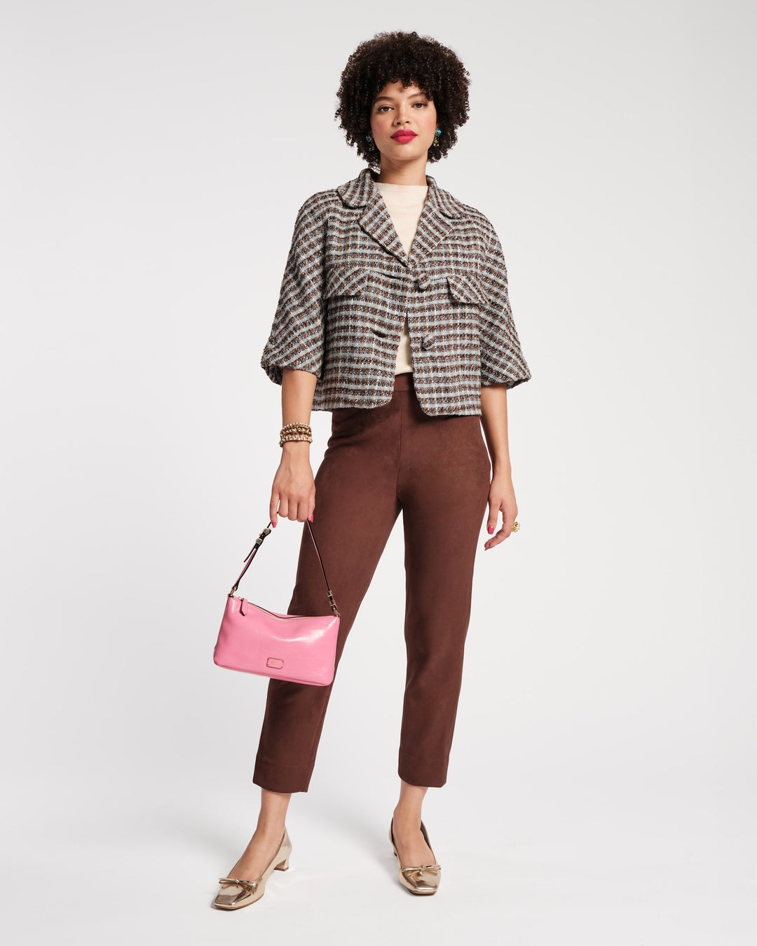 A person stands against a plain background wearing a Frances Valentine Day Jacket in Wool Park Avenue Plaid Blue Brown, a light beige top, and brown pants. They are holding a pink handbag and have on gold shoes. Their curly hair is styled naturally, and they are accessorized with bracelets.