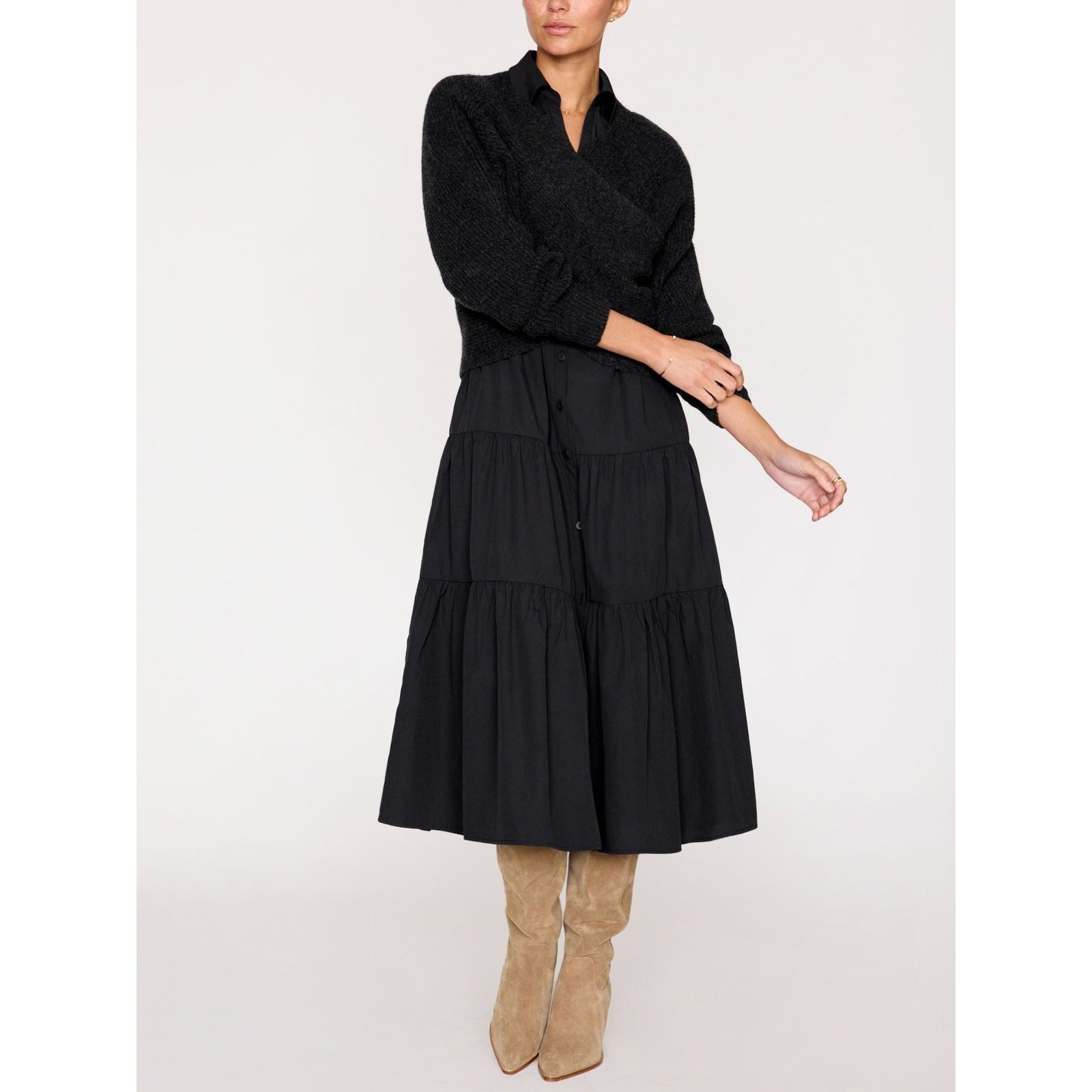 A person is seen wearing the Brochu Walker Long Sleeve Havana Dress in black. They are also wearing tall, tan suede boots. The background is plain and light-colored. The person's right arm is slightly bent, and their left hand rests on their right elbow.