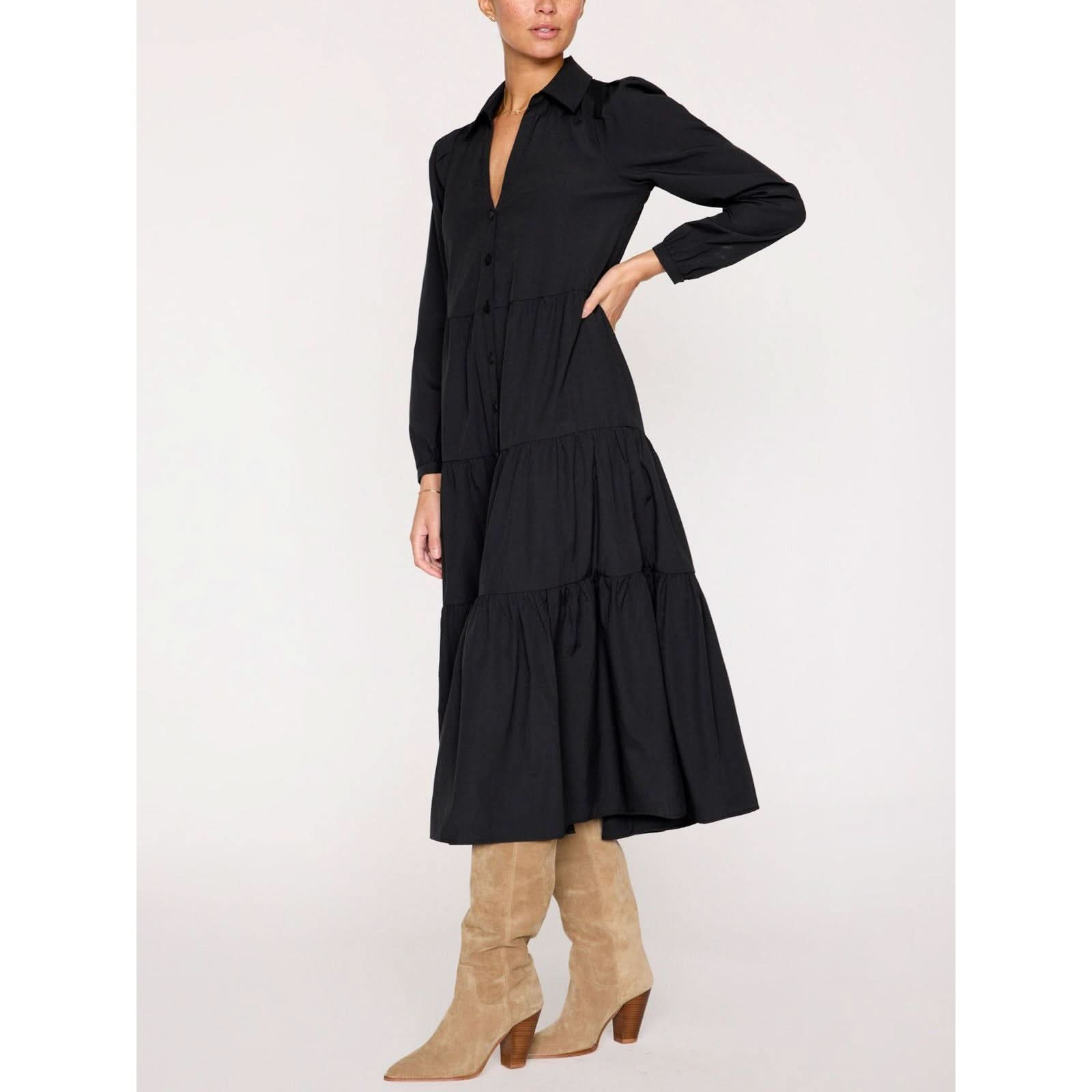 A person is standing against a plain background, wearing the Brochu Walker Long Sleeve Havana Dress in black by Brochu Walker, which features a button-up design and a tiered skirt. They are also sporting knee-high suede boots with a low heel. Their left hand is casually placed on their hip.