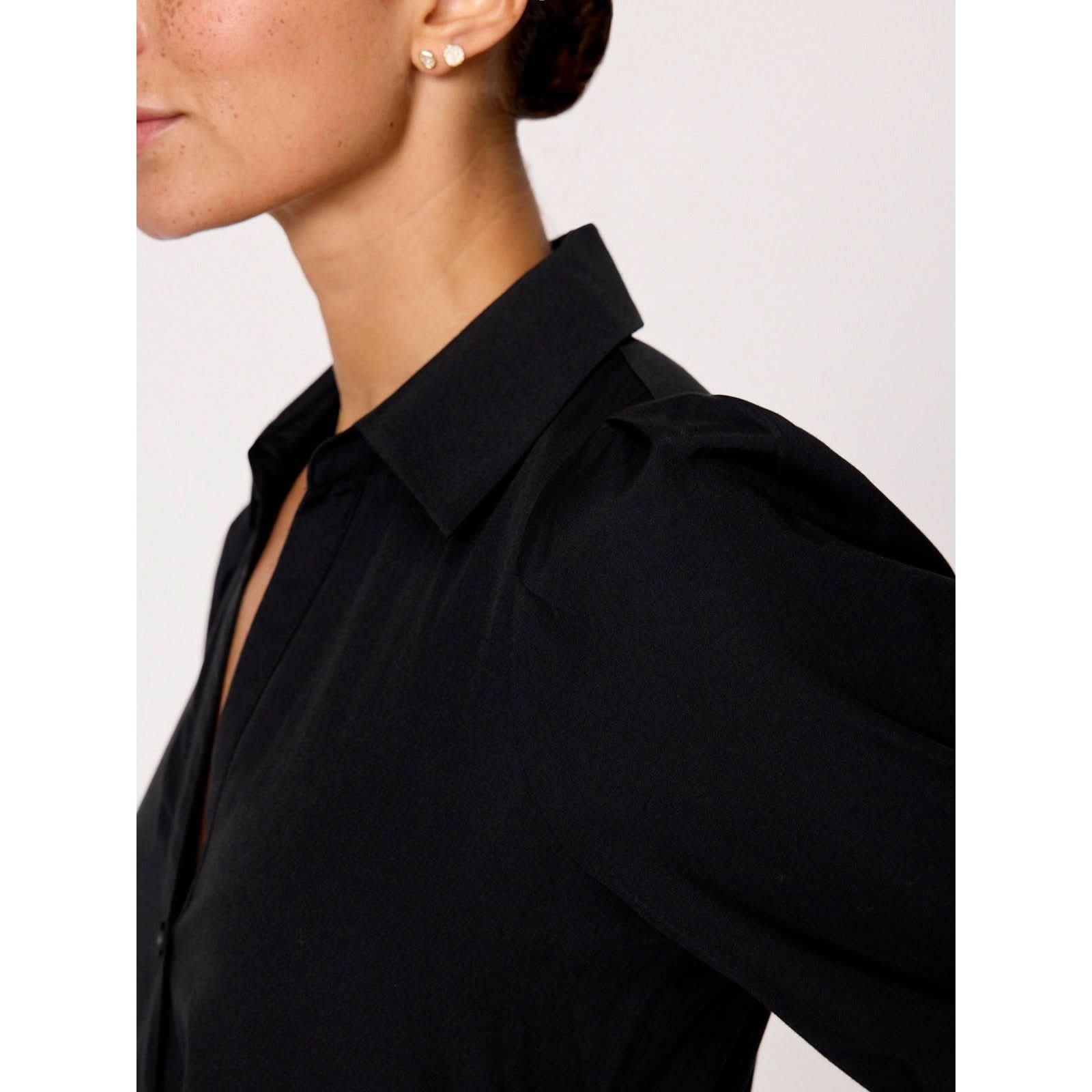 Close-up of a person wearing the Brochu Walker Long Sleeve Havana Dress in black, along with a pair of pearl stud earrings. The person has a neat bun hairstyle and is positioned against a neutral background. Only the side of their face and upper torso are visible.