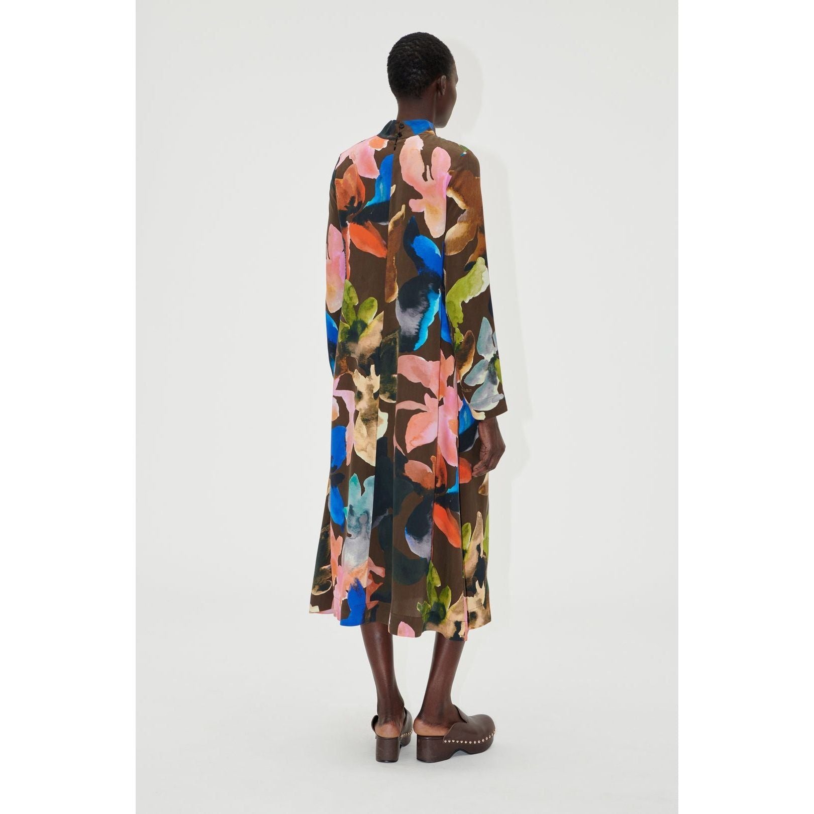 A person is turned away, showcasing the Stine Goya Long Sleeve Midi Dress - Aquarelle Garden, which displays a flowy A-line design with a vibrant abstract floral pattern in shades of green, pink, blue, and orange. The ensemble is finished with brown clogs against a simple white backdrop.