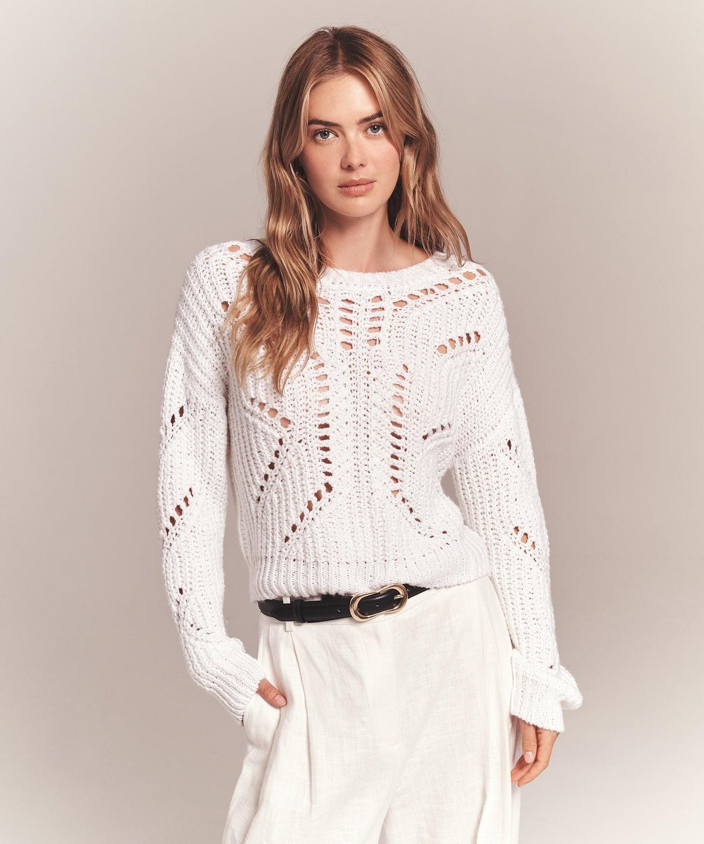 A person stands against a plain backdrop wearing a white, long-sleeved Naadam Luxe Cotton Novelty Pointelle Crew sweater featuring an intricate pattern. They have long, wavy hair and are dressed in light-colored pants with a black belt. Their hands are partially in their pockets.