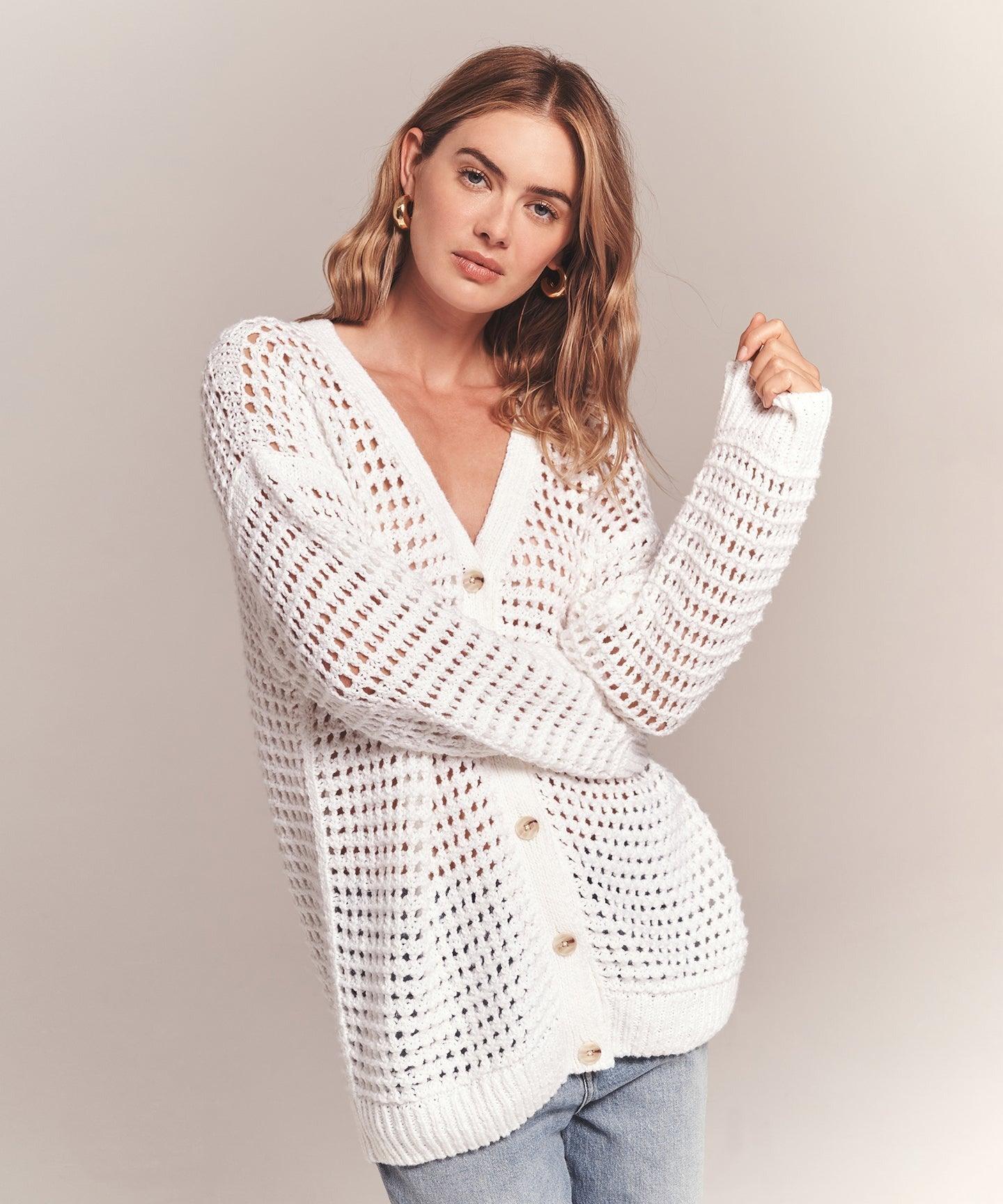 A woman with wavy hair wears a white Naadam Luxe Cotton Pointelle Cardigan, made from soft organic cotton, over a simple top and blue jeans. She stands against a plain background, gazing directly at the camera with one hand holding the edge of her cardigan.