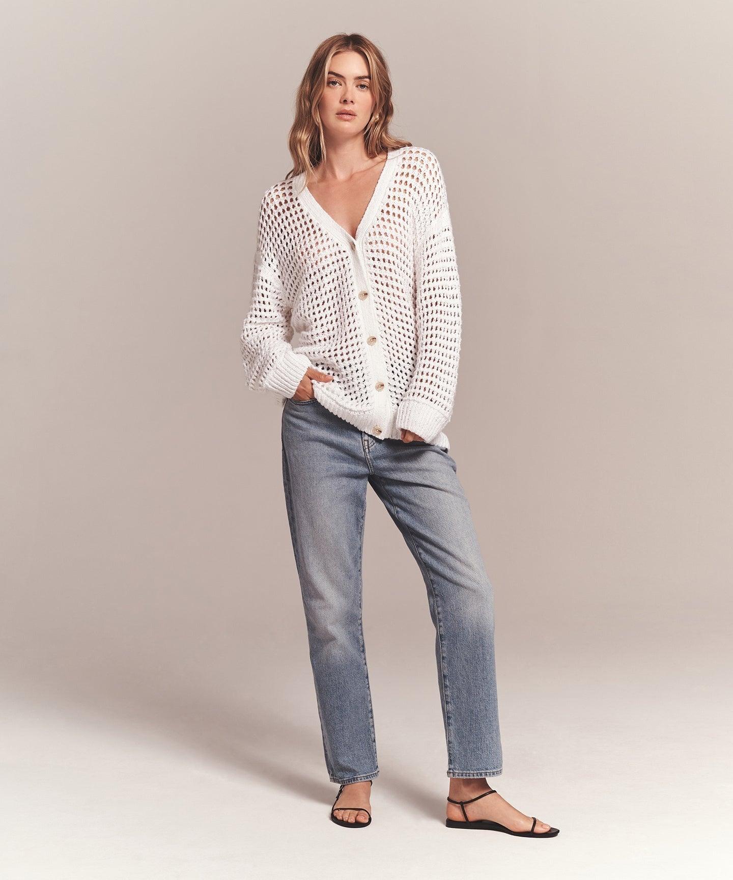 A person wearing the Naadam Luxe Cotton Pointelle Cardigan from Naadam, an oversized white sweater with chunky pointelle stitching, over a beige top, blue jeans, and black strappy sandals stands with one hand in their pocket against a plain, light-colored background. They have long hair and a relaxed pose.