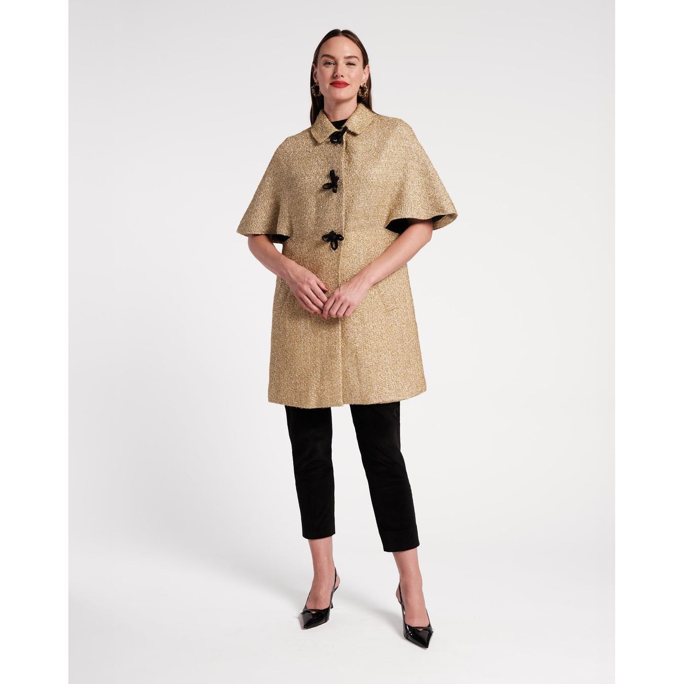 In a studio space with a plain white background, someone is modeling the Frances Valentine Marvelous Jacket Rockefeller Gold. This stylish jacket features short capelet sleeves and black toggle buttons, paired with black pants and sleek black heels.