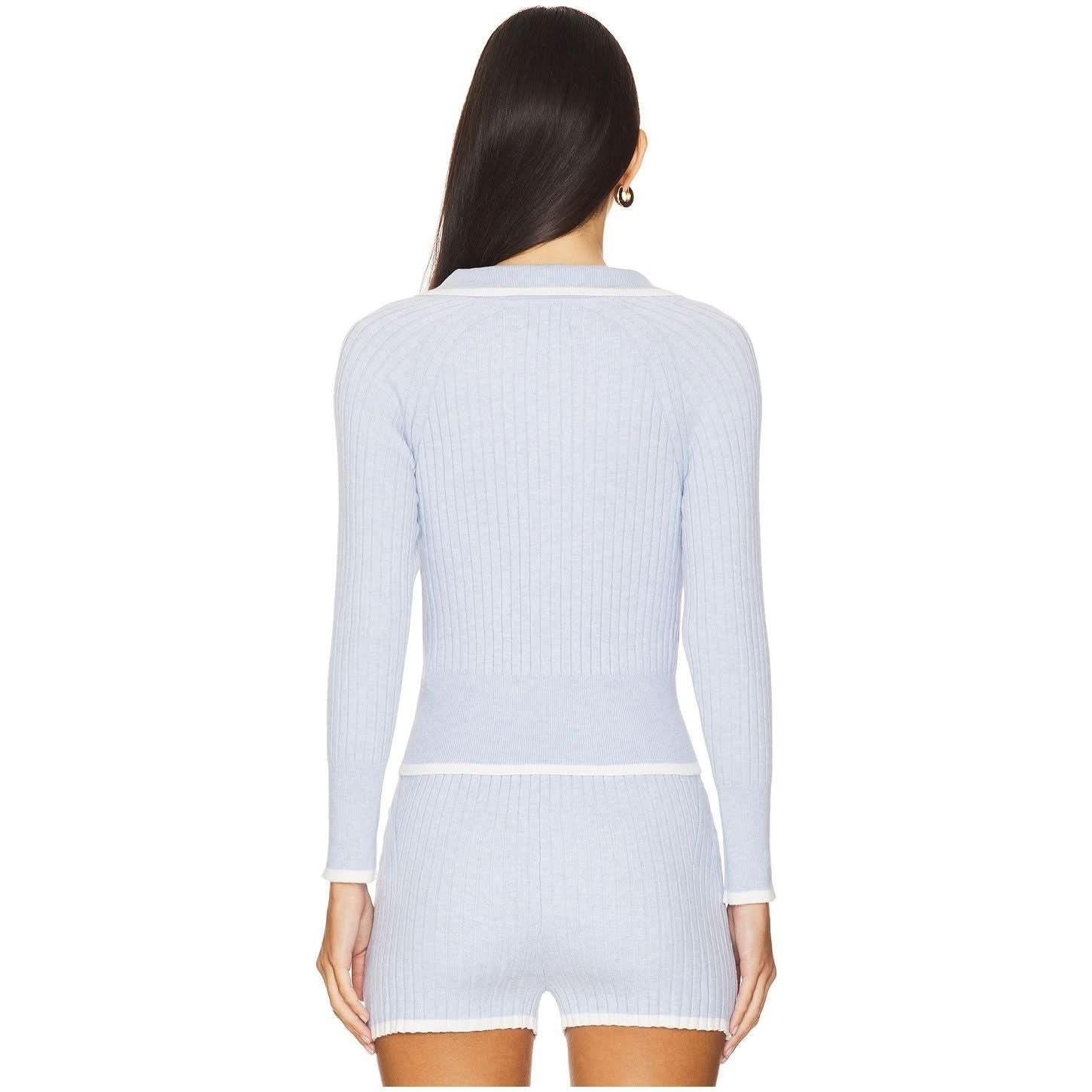 A woman with long dark hair is seen from the back wearing a Matthew Bruch Rib Knit Light Blue Cropped Cardigan and matching shorts made from recycled nylon. The outfit features white trim at the cuffs, hem, and waistband.