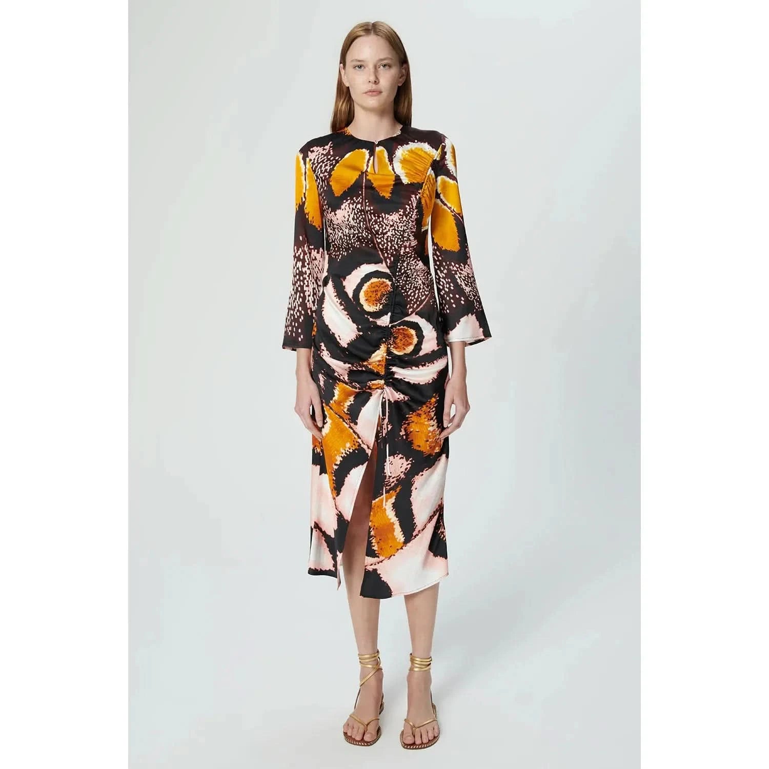 A woman stands against a plain background wearing the Chufy Orchid Maxi Dress by Chufy, made from sustainable fabric and featuring a colorful, abstract print with long sleeves and a ruched front. She has straight, mid-length brown hair and is wearing strappy, nude sandals.