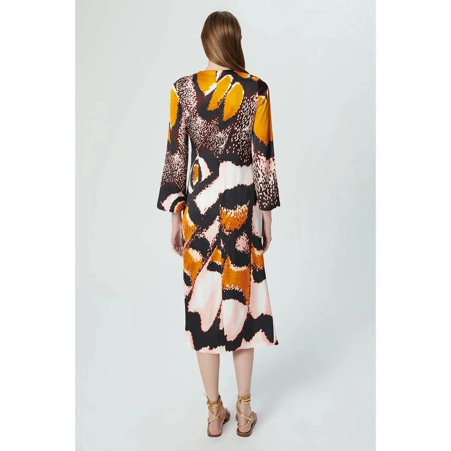 A woman with long, straight brown hair stands with her back to the camera, wearing a Chufy Orchid Maxi Dress made from sustainable fabric. The colorful, patterned dress features bold abstract designs in shades of yellow, orange, black, and white. She completes the look with strappy sandals.