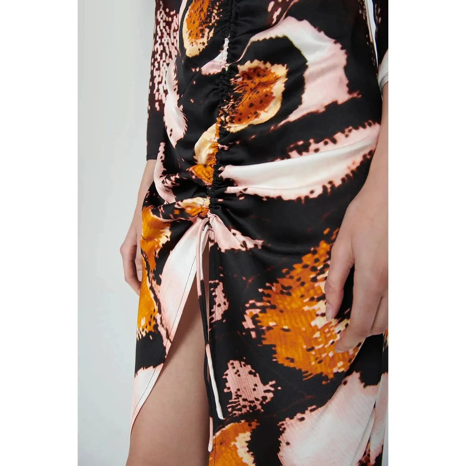 Close-up of a person wearing the Chufy Orchid Maxi Dress by Chufy, showcasing its abstract, colorful pattern in shades of black, pink, orange, and white. Crafted from sustainable fabric, the dress features ruched detailing at the waist and a high slit. The person's arm is resting at their side.