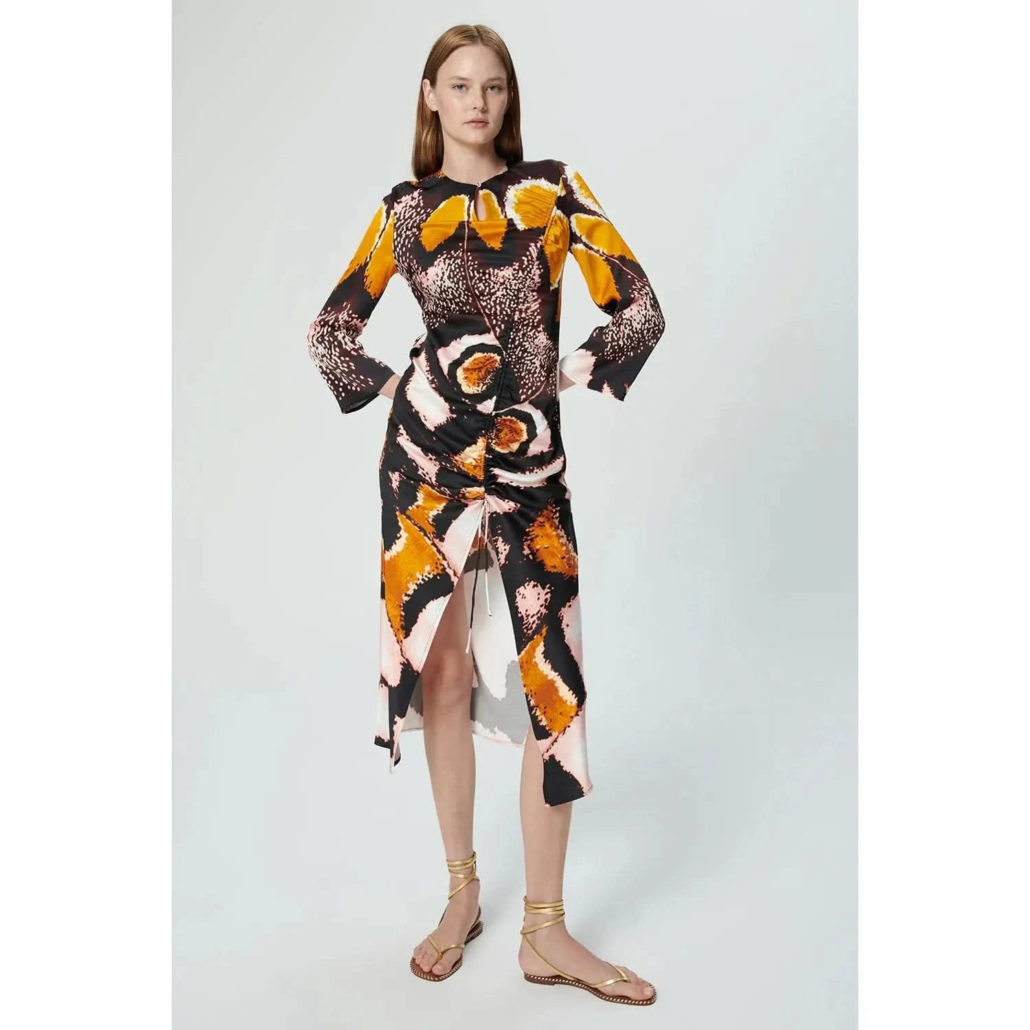 A model is wearing the Chufy Orchid Maxi Dress, a long-sleeved, multicolored midi dress by Chufy. Crafted from sustainable fabric, it showcases a bold abstract print in orange, black, beige, and white hues. The dress features a ruched front with a tie detail and a slit. She pairs it with flat, strappy sandals against a plain background.