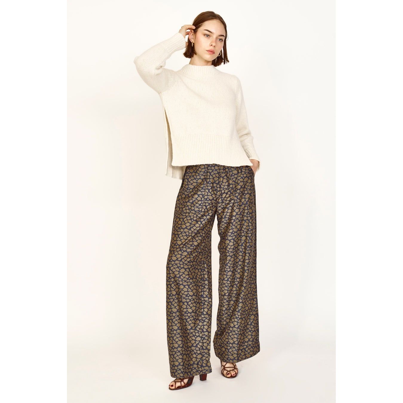 A person poses against a neutral background, wearing a cream-colored silk sweater and the Mirth Tivot Pant Navy Lilypad, which feature a hand screen printed, wide-leg design. They have one hand in their hair and look confidently toward the camera.