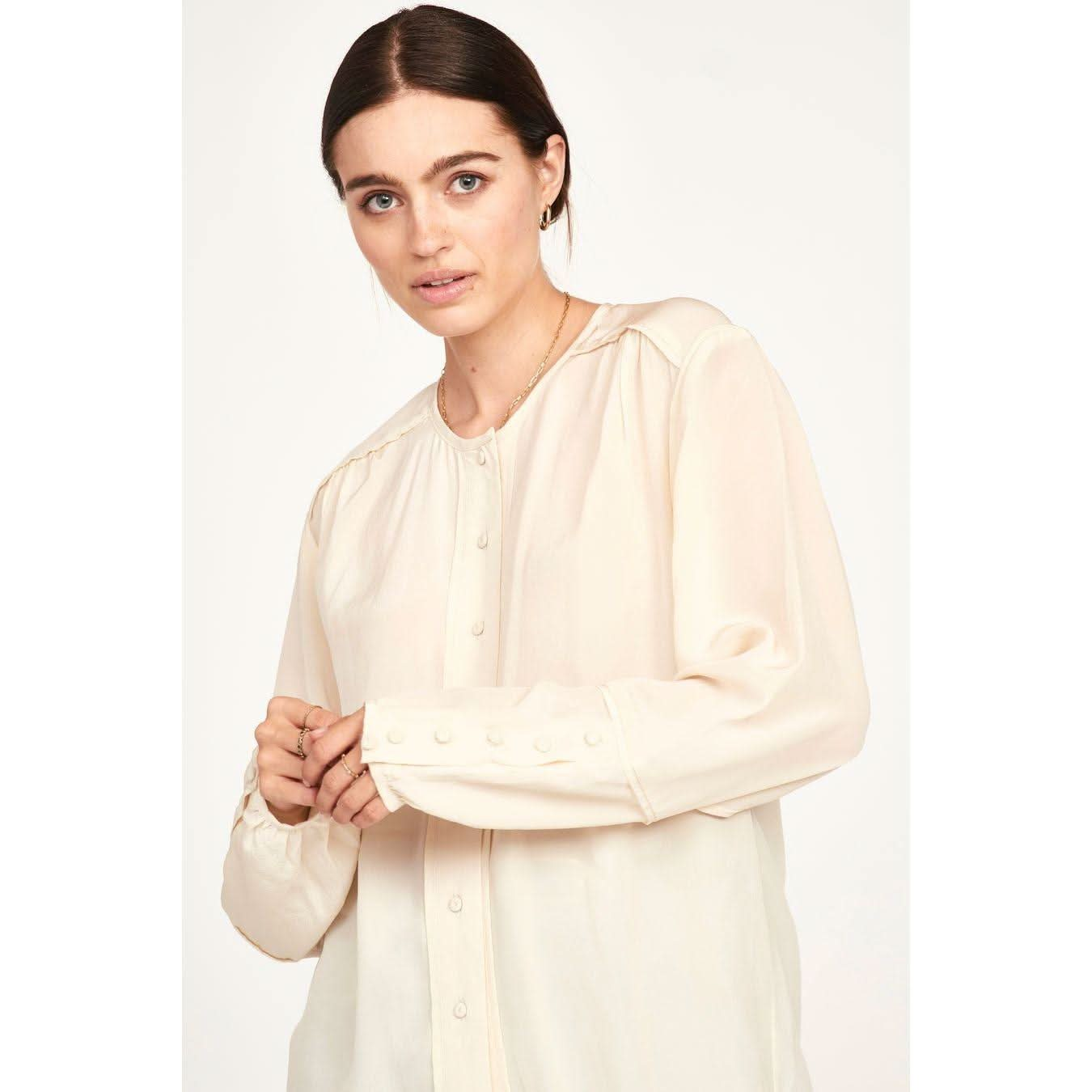 A person with long dark hair, wearing the Mirth Dakota Blouse Ivory by Mirth, which features an oversized fit in light beige silk with buttoned cuffs, stands facing the camera against a plain background. They have a neutral expression and are holding their sleeve with one hand.