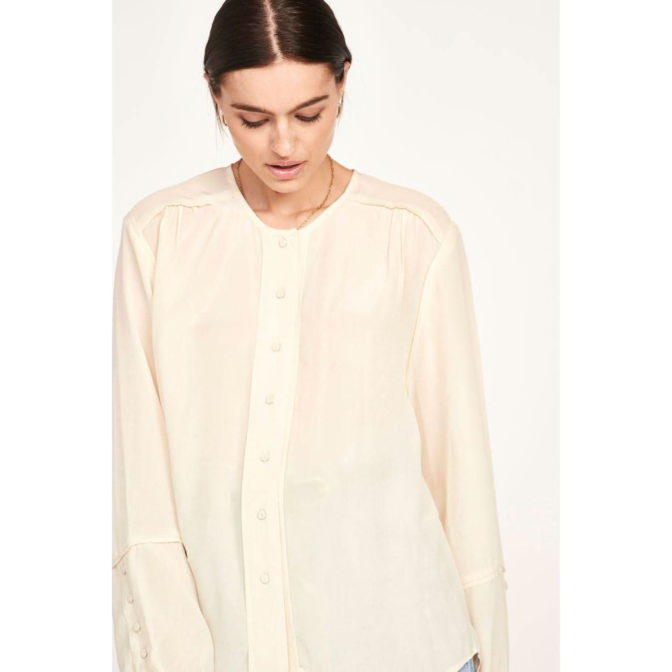 A person with dark hair tied back is wearing the Mirth Dakota Blouse in ivory, a cream-colored, sheer silk blouse featuring an oversized fit. They are looking down slightly against a plain, light background.