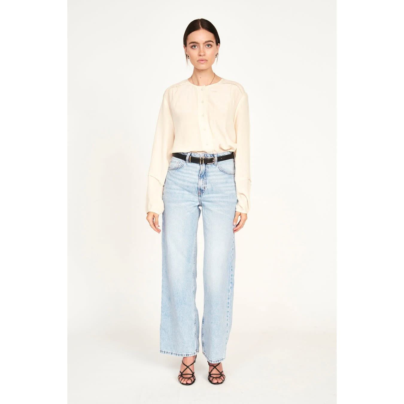 Standing against a simple backdrop, the individual dons the Mirth Dakota Blouse in Ivory, paired with light blue wide-leg jeans and black strappy sandals. Their long dark hair is pulled back, and they wear a neutral expression.