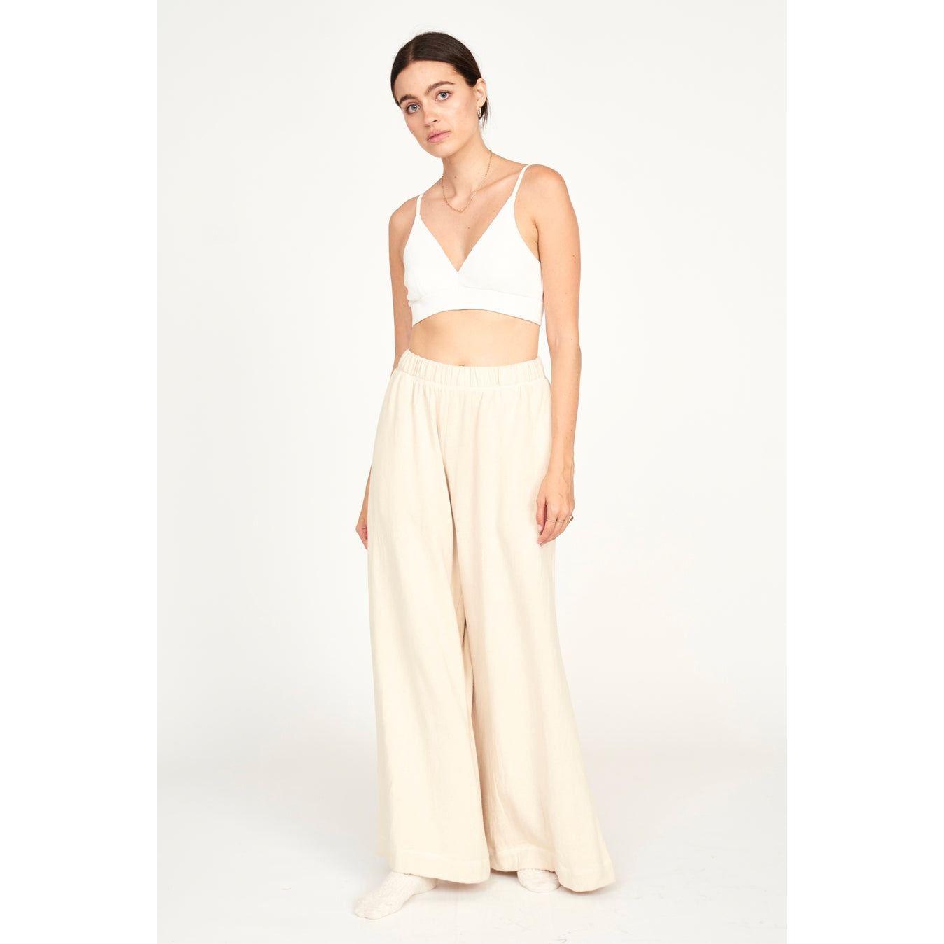 A woman stands in a relaxed pose against a plain white background, wearing the Mirth Lounge Pant in Dove—a loose, beige high-waisted pant made from sustainable fabric—and a white crop top. She has long dark hair and minimal makeup.