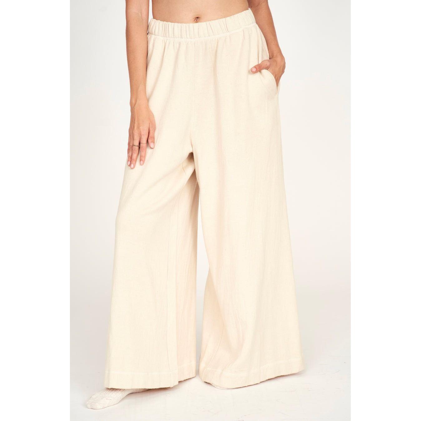 A person wearing the Mirth Lounge Pant in Dove, which are light beige wide-legged pants crafted from regenerative cotton, is seen with a hand in the pocket. These naturally dyed pants feature an elastic waistband and are photographed against a plain background, emphasizing their sustainable production by Mirth.