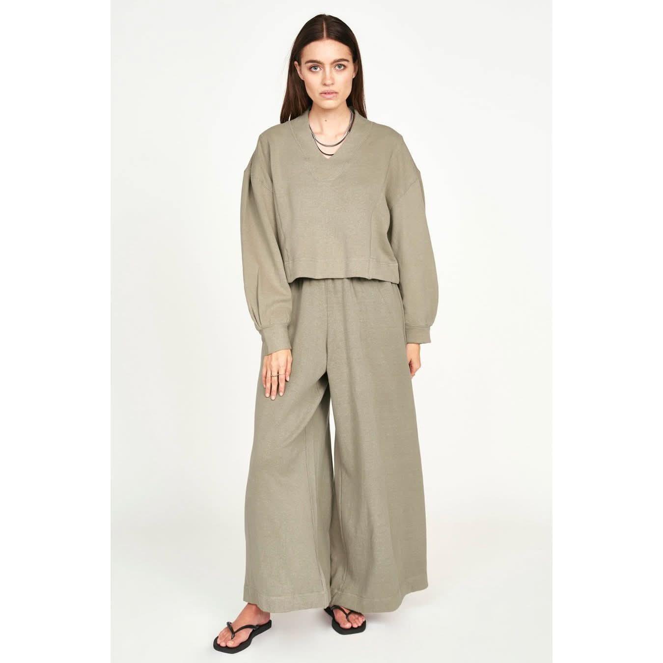 A person with long hair is wearing a loose-fitting, sage green Mirth Canyon Sweatshirt in Sagebrush made from sustainable fabric, paired with wide-leg pants. They are also wearing black flip-flops and standing against a plain white background, with their hands relaxed by their sides.