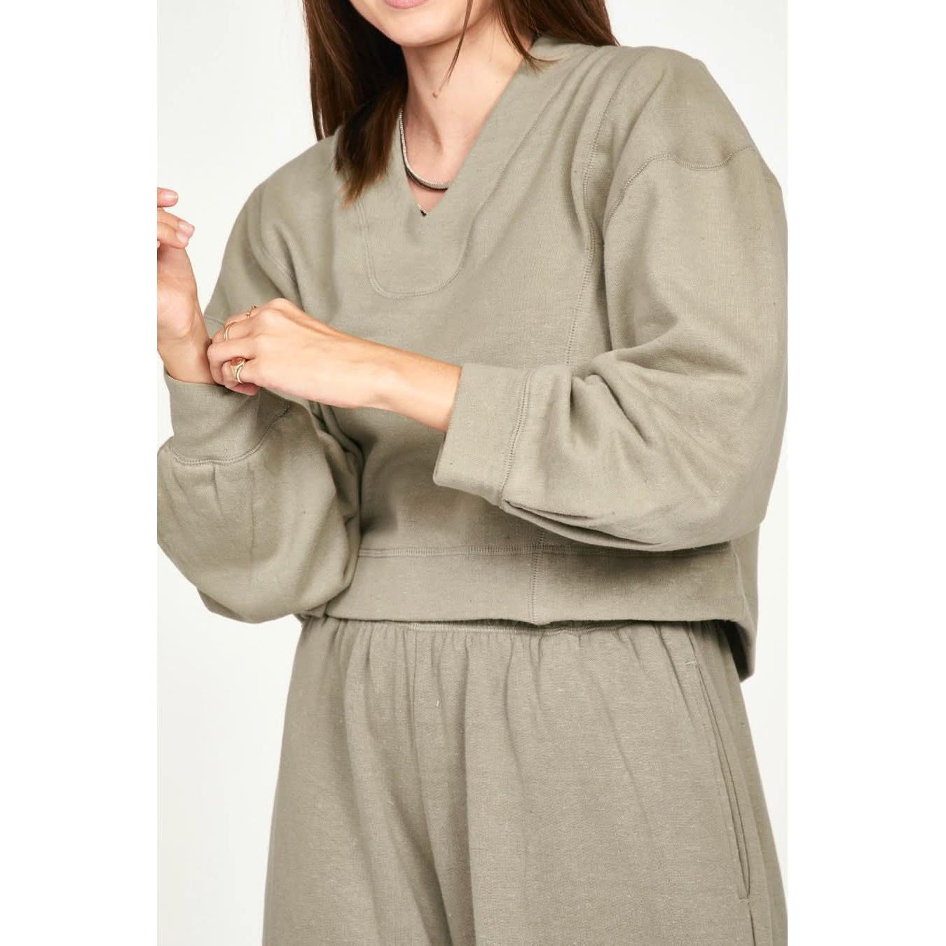 A person wearing a cozy, oversized Mirth Canyon Sweatshirt in Sagebrush and matching jogger pants made from sustainable fabric. The Mirth sweatshirt, crafted from regenerative cotton, features long sleeves and a V-neckline. The individual's face is not visible, and they are accessorizing with a simple necklace and a ring.