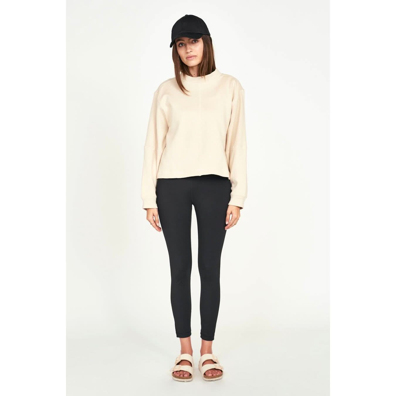 A person wearing a black cap, the Mirth Cordoba Sweatshirt in Dove made from regenerative cotton, black leggings, and white sandals stands against a plain background. Their hands are at their sides, and they are looking forward.