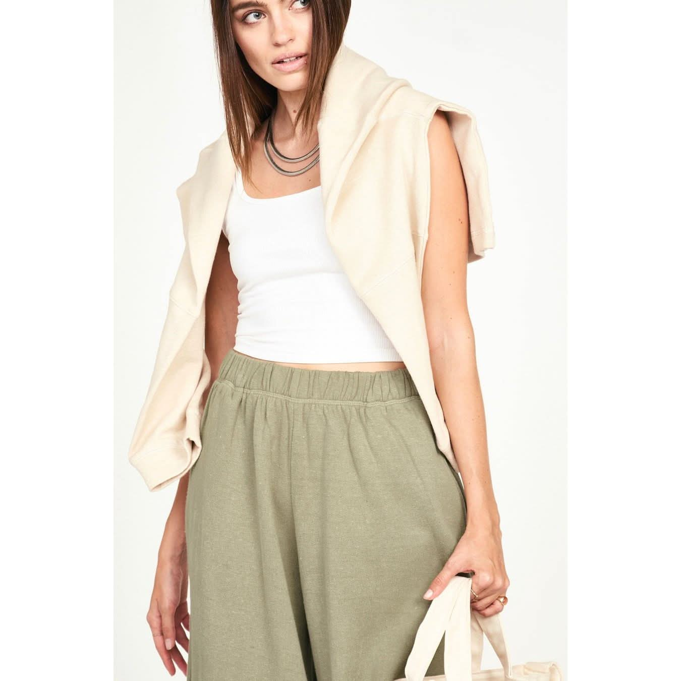 A person with shoulder-length brown hair wears a white tank top and olive green pants made of sustainable fabric, accessorized with the Mirth Cordoba Sweatshirt in Dove draped over their shoulders. They carry a beige bag and wear a layered necklace.