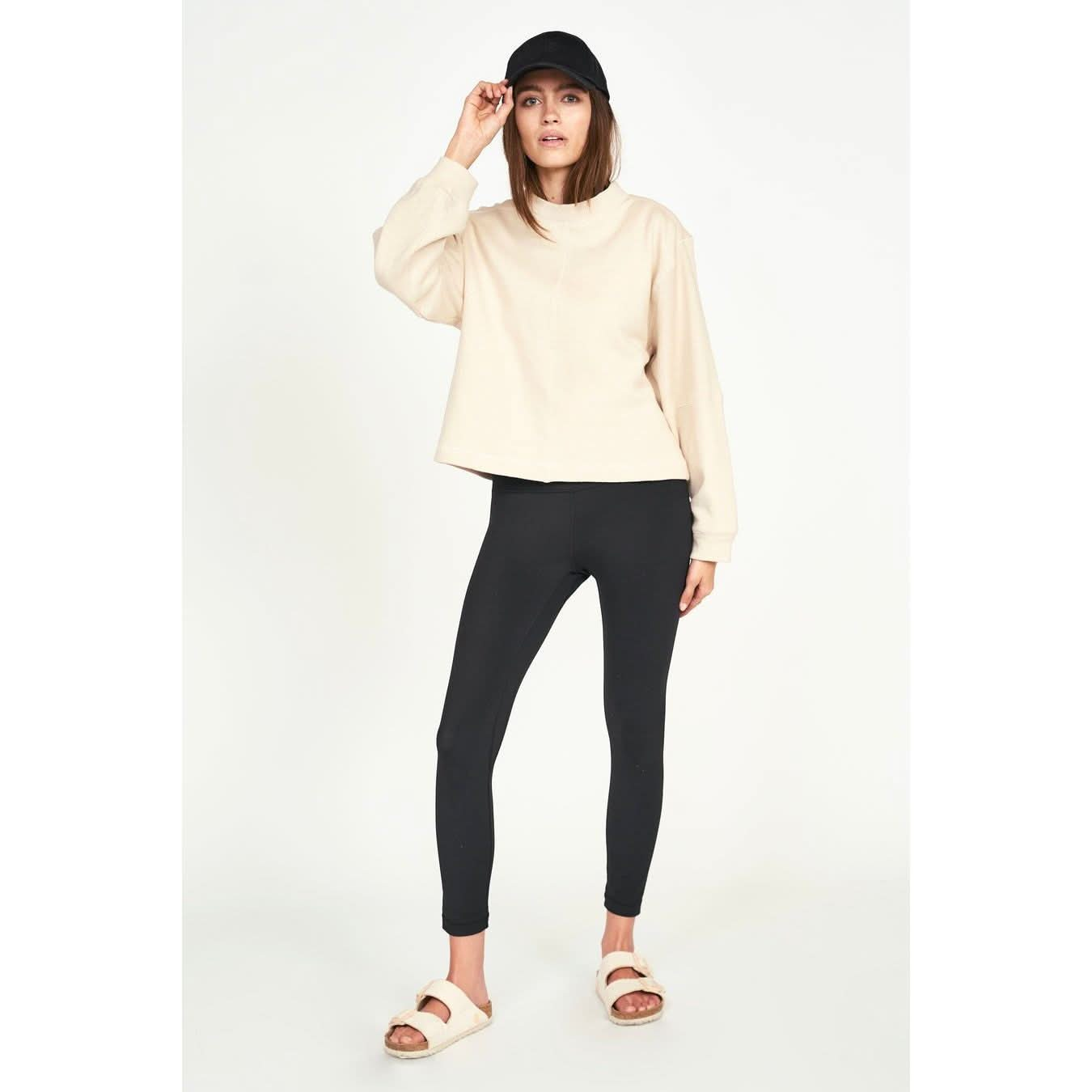 Against a plain background, a person wears the Mirth Cordoba Sweatshirt in Dove, complemented by black leggings and white sandals. They sport a black cap and strike a relaxed pose, with one hand touching the cap as they look at the camera.