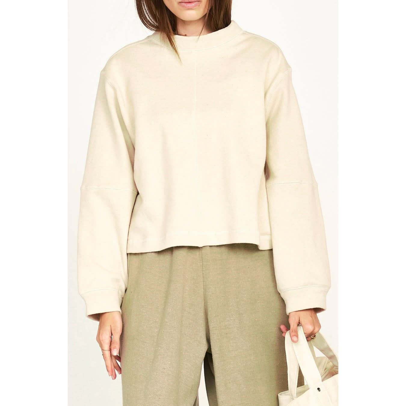 A person is wearing the Mirth Cordoba Sweatshirt in Dove, an oversized beige sweatshirt with long sleeves, paired with high-waisted, wide-leg beige pants made from naturally dyed, sustainable fabric. They are holding a matching beige tote bag in their left hand and their head is partially out of the frame.