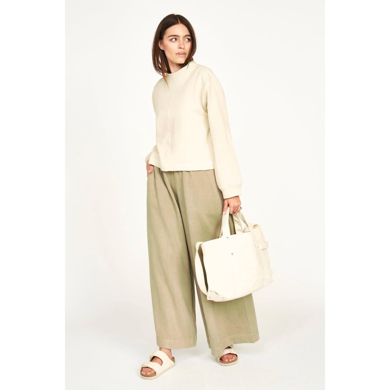 A woman dressed in the Mirth Cordoba Sweatshirt in Dove, made from regenerative cotton, complements her outfit with wide-legged khaki pants and a matching cream tote bag. She stands confidently with one hand in her pocket against a plain backdrop, showcasing her white sandals, each item reflecting the elegance of sustainable production.