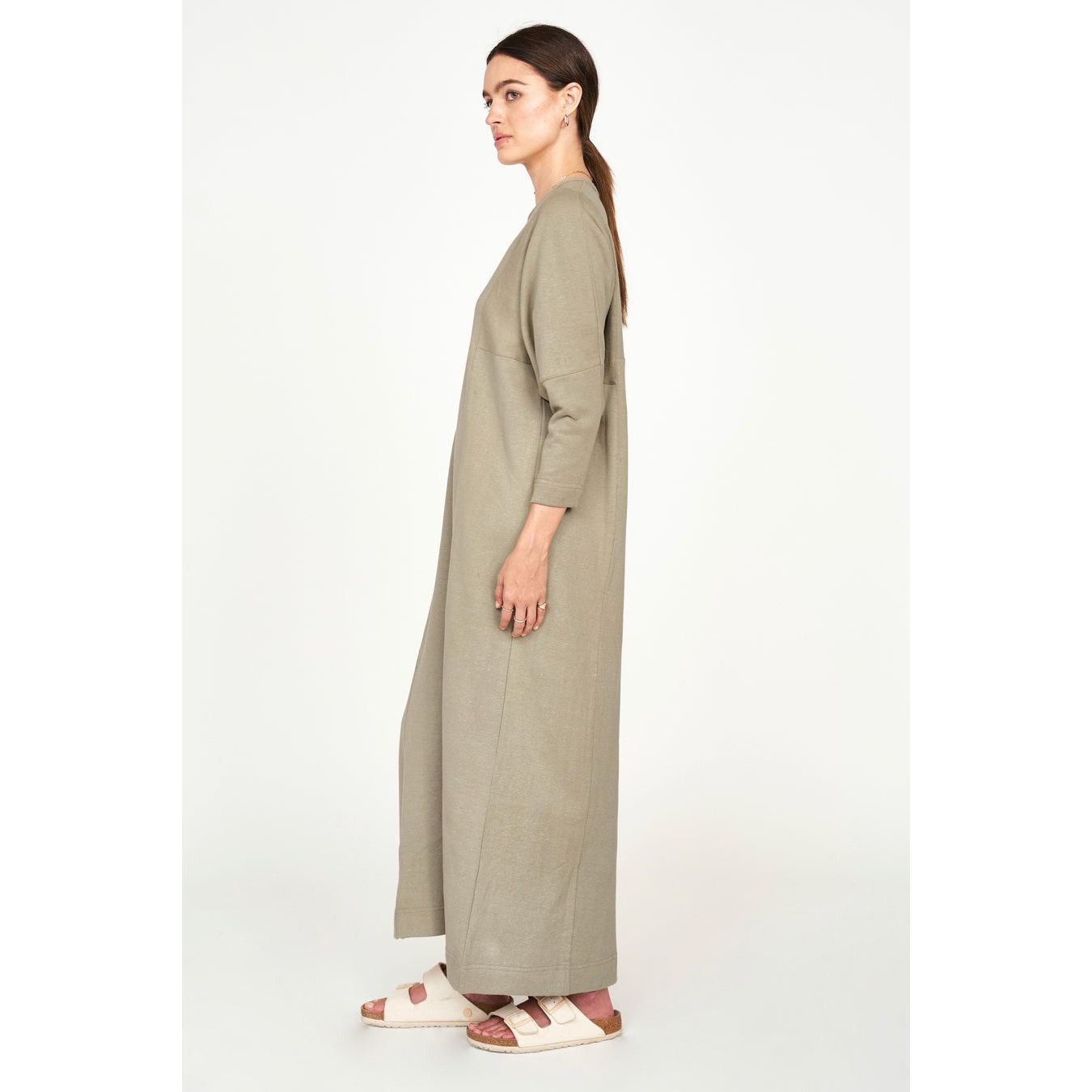 A person wearing the Mirth Curacao Lounge Dress in Sagebrush, crafted from sustainable fabric, poses in profile against a plain background. Their long dark hair is tied back, and they are wearing white sandals.