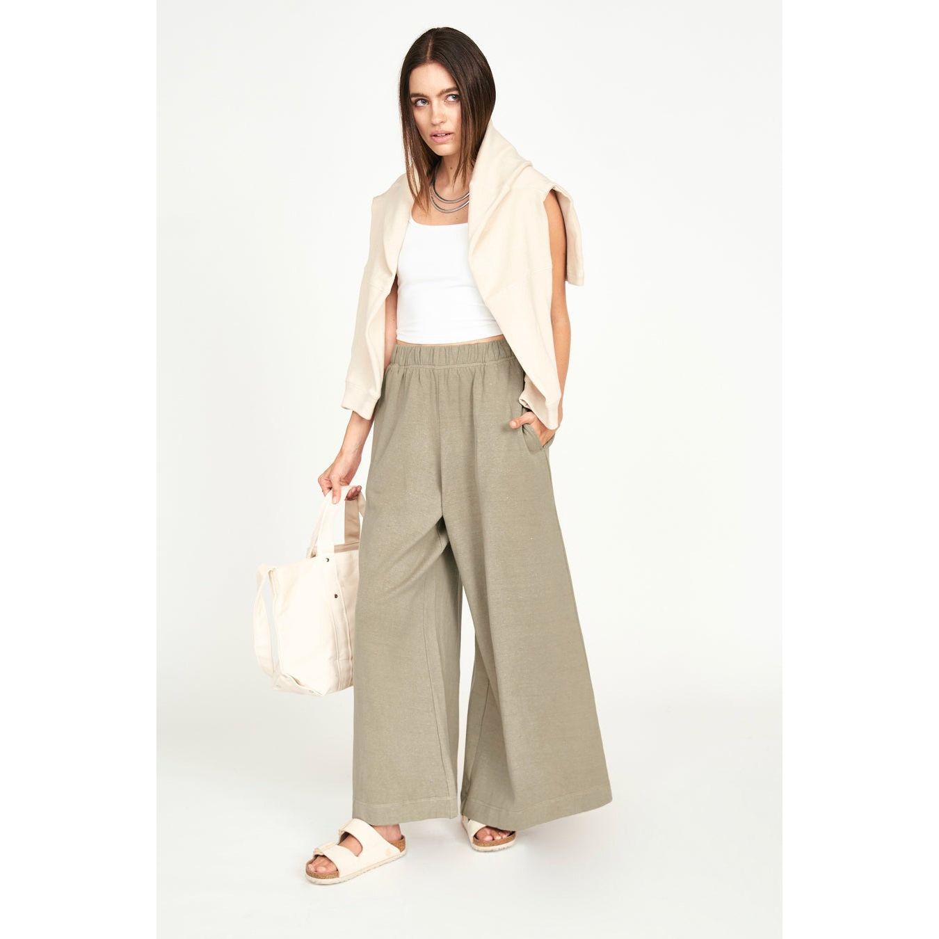 A woman stands against a plain background wearing a white tank top, the Mirth Lounge Pant in Sagebrush made from regenerative cotton, and a light sweater draped over her shoulders. She accessorizes with beige sandals, a beige bag in her left hand, and a layered necklace.