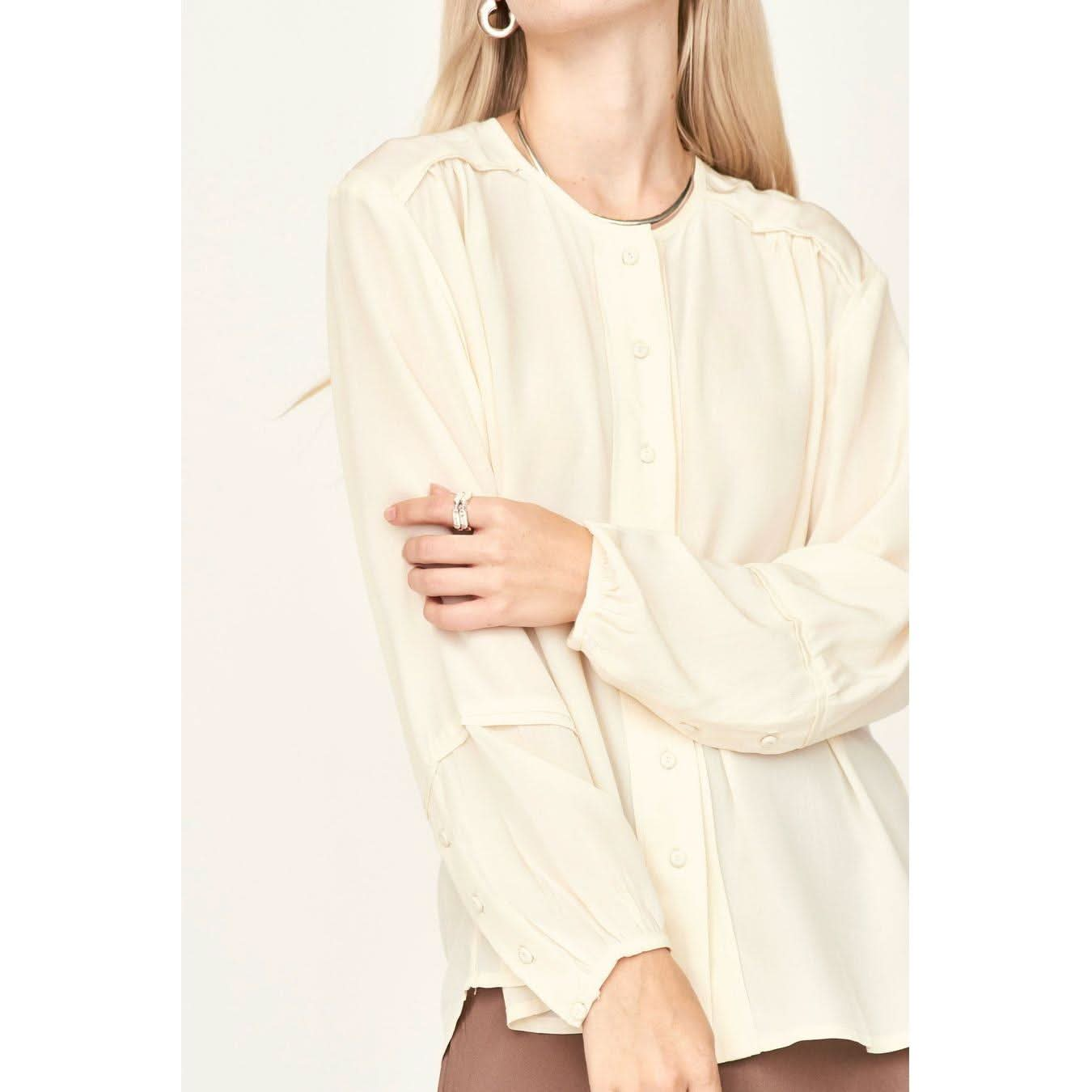 A woman with long blonde hair is wearing the Mirth Dakota Blouse in ivory, paired with brown pants. The slightly oversized fit of her sleeves adds a touch of elegance as she crosses one hand over her body. Hoop earrings and rings complete her look against a sheer, light-colored background.