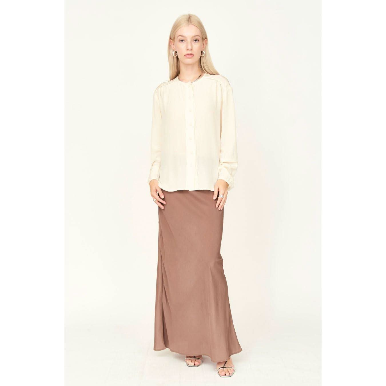 A woman in the Mirth Dakota Blouse in Ivory and a long brown skirt stands against a light background. Her straight hair frames her neutral expression. She complements the ensemble with simple jewelry and sandals, adding a touch of elegance to the understated look.