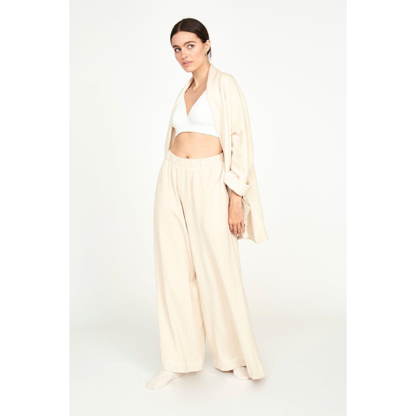 A woman casually stands against a plain white background, wearing the Mirth Lounge Pant in Dove, a loose, cream-colored outfit made from sustainable fabric. The ensemble consists of wide-legged pants, a draped open cardigan, and a white bralette. Her dark hair is pulled back.