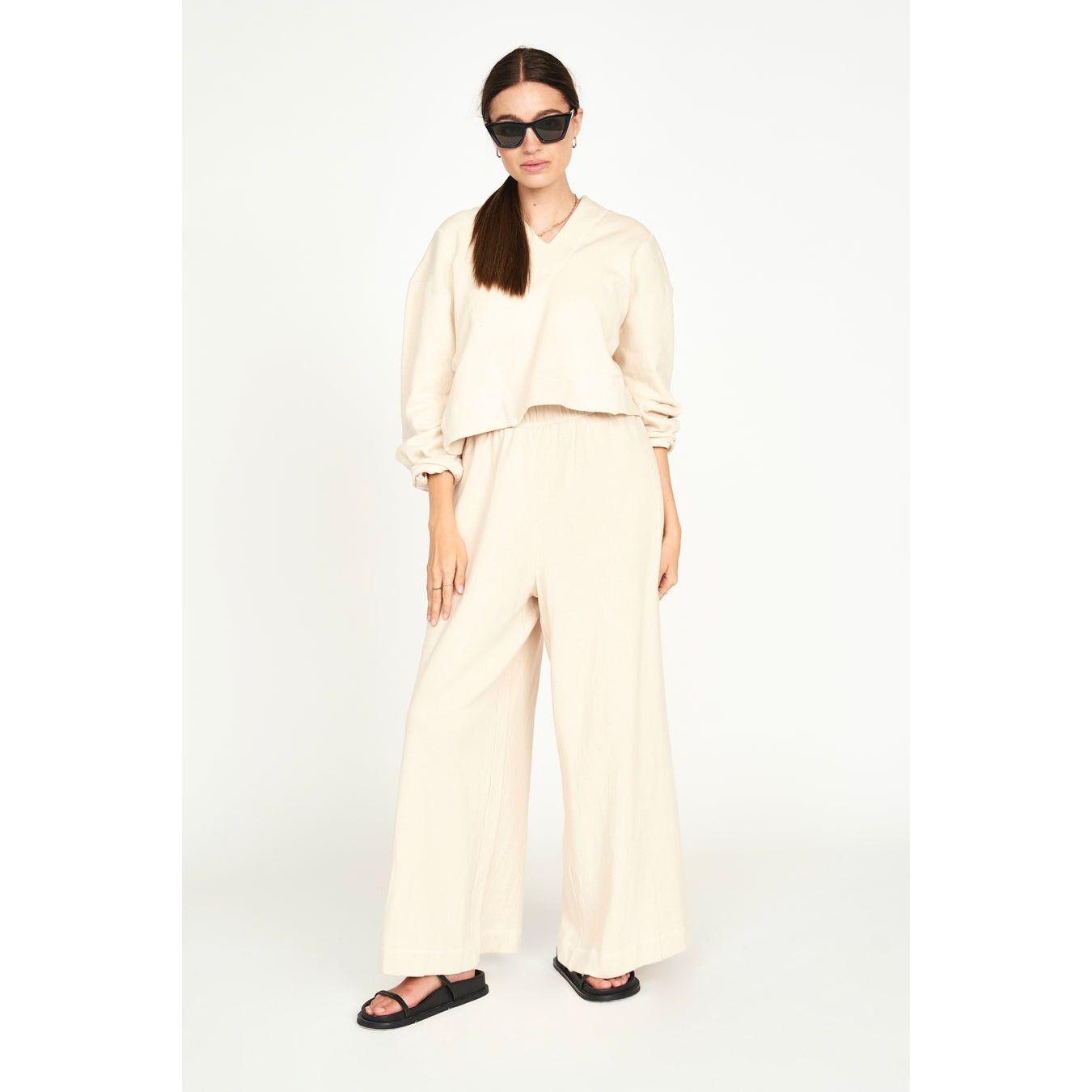 A woman stands confidently, wearing the Mirth Lounge Pant in Dove along with a matching long-sleeve top and oversized sunglasses. The naturally dyed, regenerative cotton outfit complements her dark hair, which is tied back, as she sports black sandals. She poses against a plain white background.