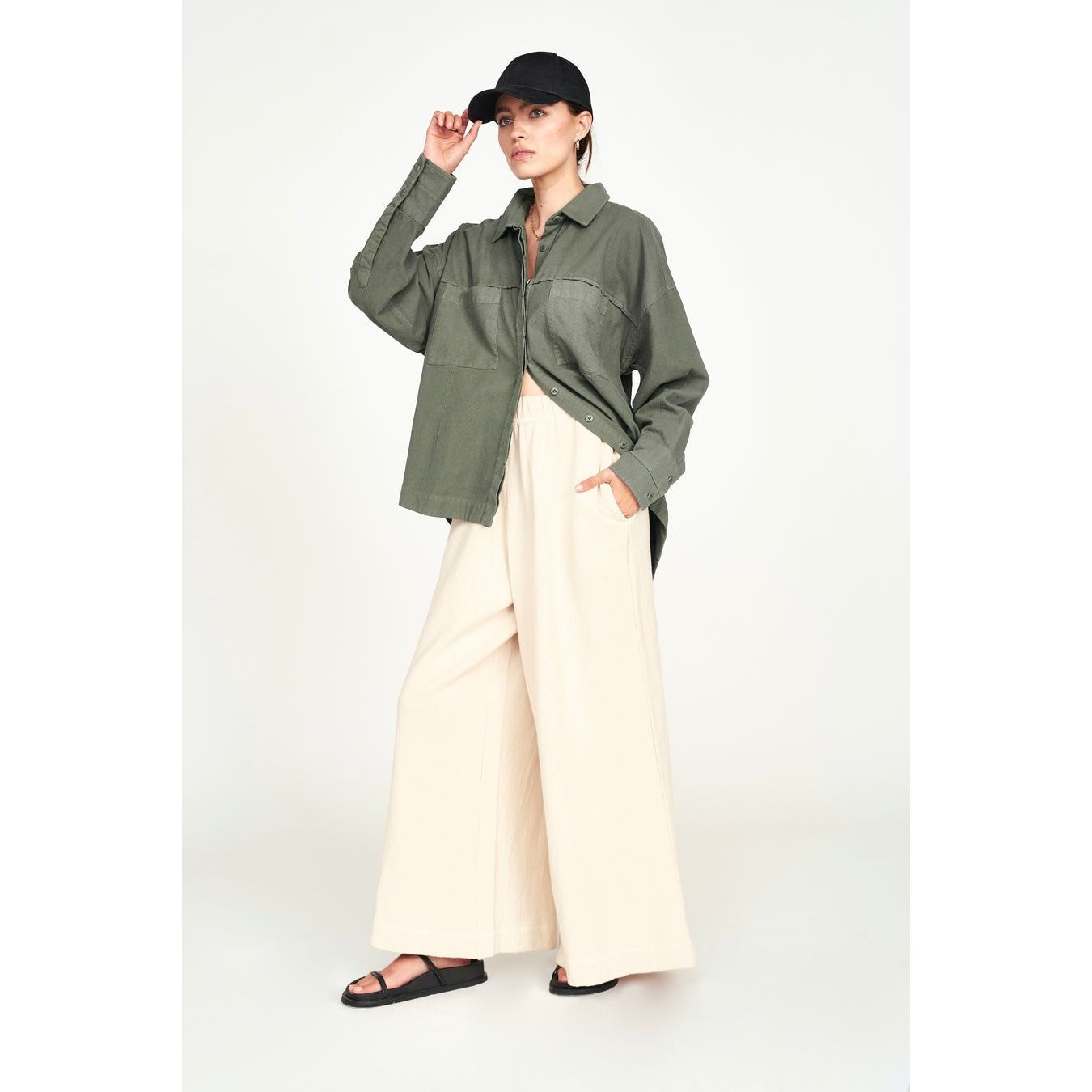 A person dressed in a dark cap, an oversized naturally dyed green button-up shirt, and the Mirth Lounge Pant in Dove from Mirth stands against a plain background. They gaze to the side, adjusting their cap with one hand.