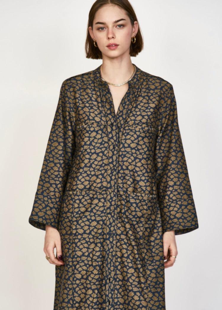 A person with shoulder-length hair is wearing a long-sleeved, patterned Mirth Malmo Dress in Navy Lilypad, made in India. The hand screen-printed design features an intricate, repeating pattern in a mix of brown and dark colors. They are standing against a plain background.