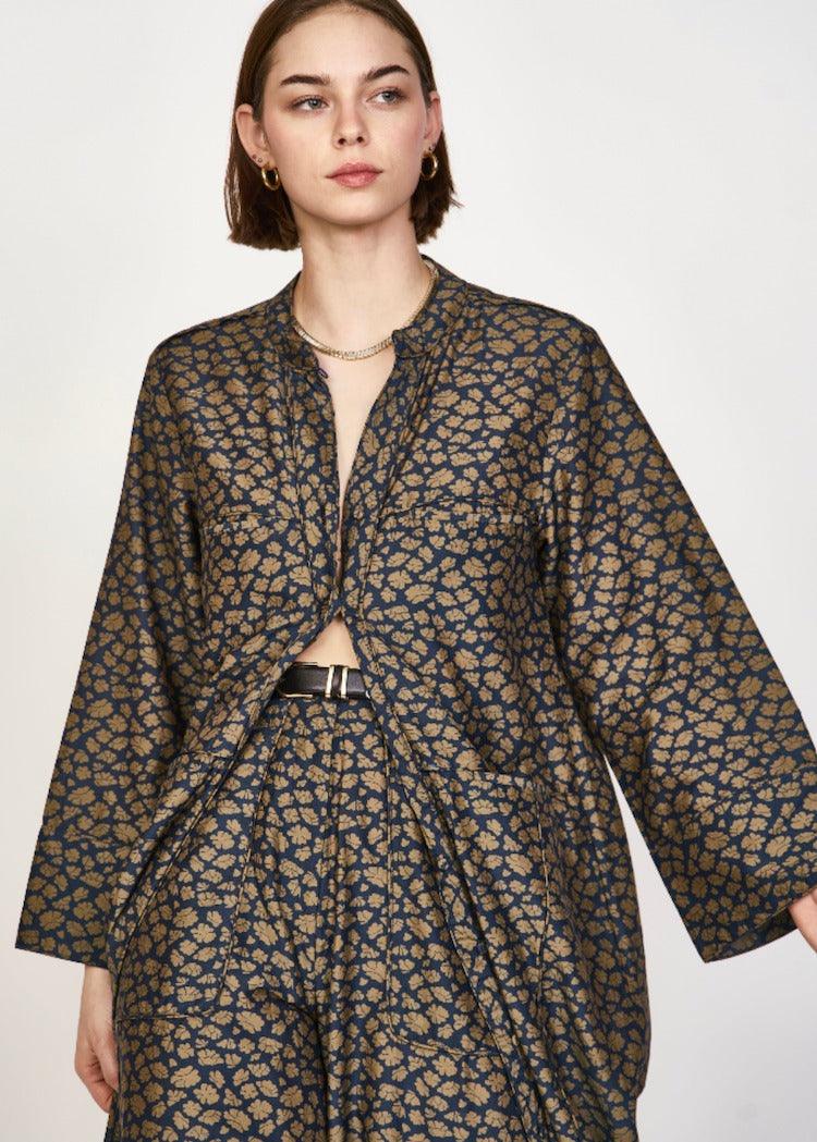 A woman with short brown hair is wearing the Mirth Malmo Dress in Navy Lilypad, which features a dark intricate gold pattern. The hand screen-printed dress, made in India, has an open front that reveals part of her torso. She accessorizes with understated jewelry.