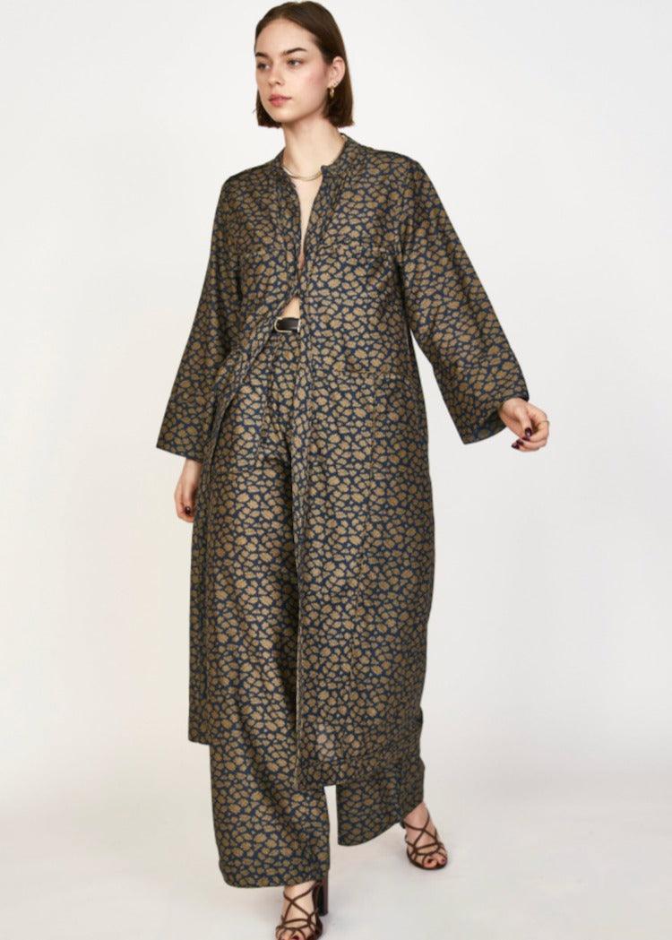 A model wearing the Mirth Malmo Dress in Navy Lilypad, featuring a long, patterned open-front coat and wide-leg trousers. The fabric, hand screen printed and Made in India, showcases a detailed blue and brown geometric design. The model is posing against a plain white background, accessorized with dark strappy sandals.