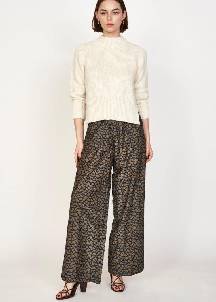 A person with straight, shoulder-length hair stands against a plain background. They are wearing a cream-colored, silk sweater with long sleeves and a high neckline, paired with the wide-legged Mirth Tivot Pant Navy Lilypad from Mirth, featuring hand screen-printed designs in shades of dark brown and black. They complete their look with open-toe heeled sandals.